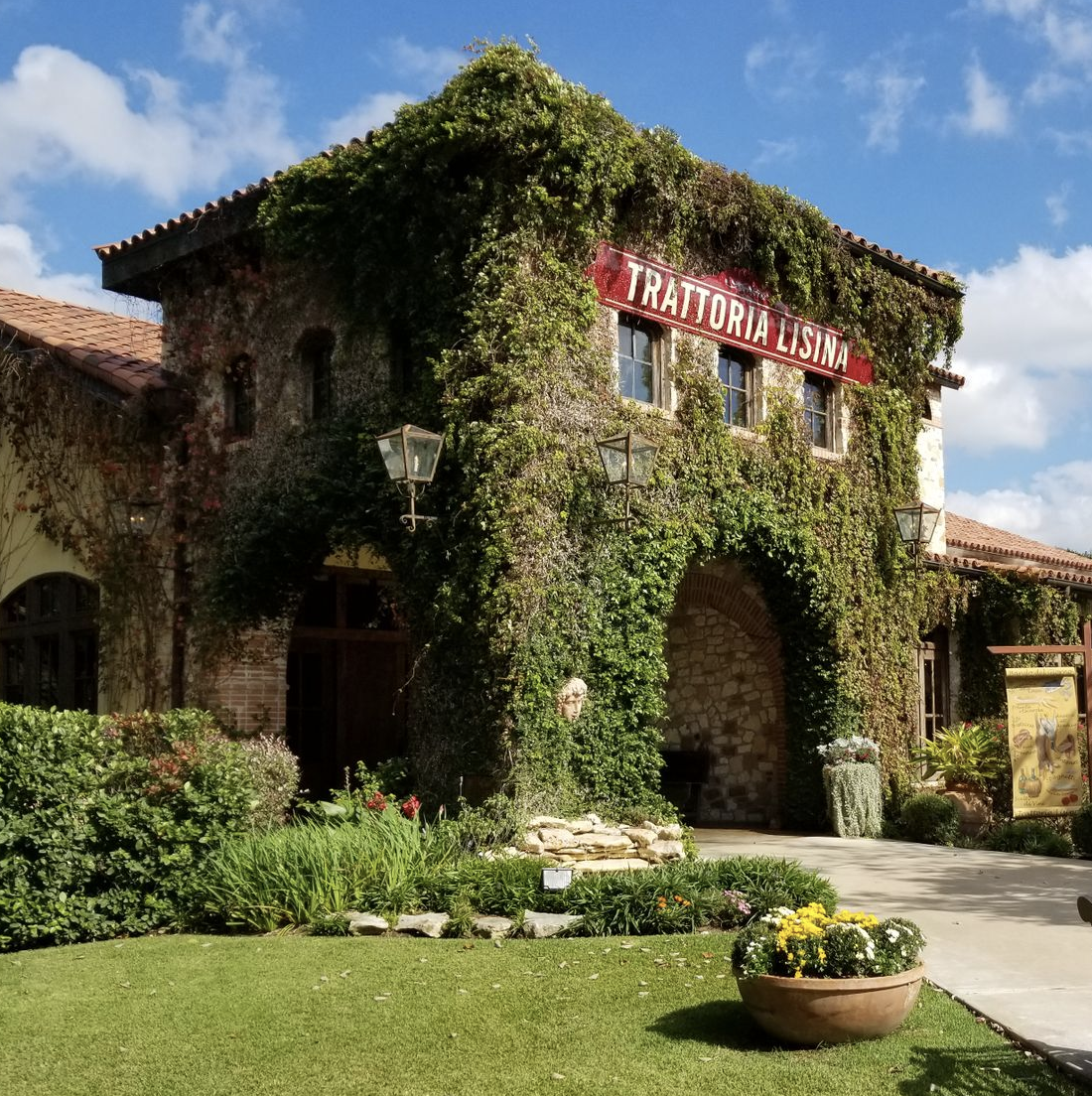 A rustic building covered in green ivy with a red sign reading 'Trattoria Lisina' on the front. There is a garden with colorful flowers and a stone pathway leading to an arched entrance, with sky and clouds overhead.