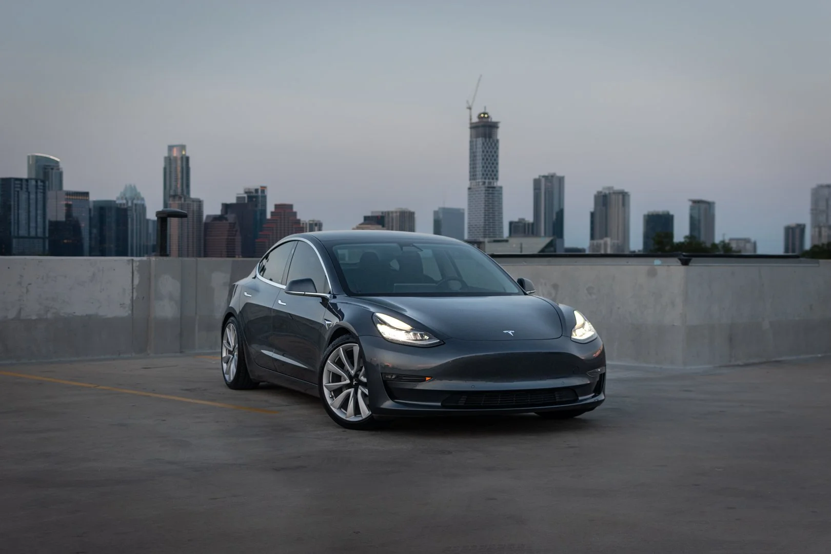 A sleek black Tesla Model 3 parked on the top level of a parking garage with a city skyline, including high-rise buildings and a construction crane, in the background.