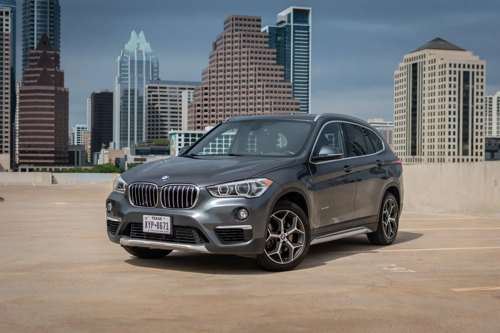 A gray BMW X1 parked in an outdoor parking lot with a city skyline of tall buildings in the background.