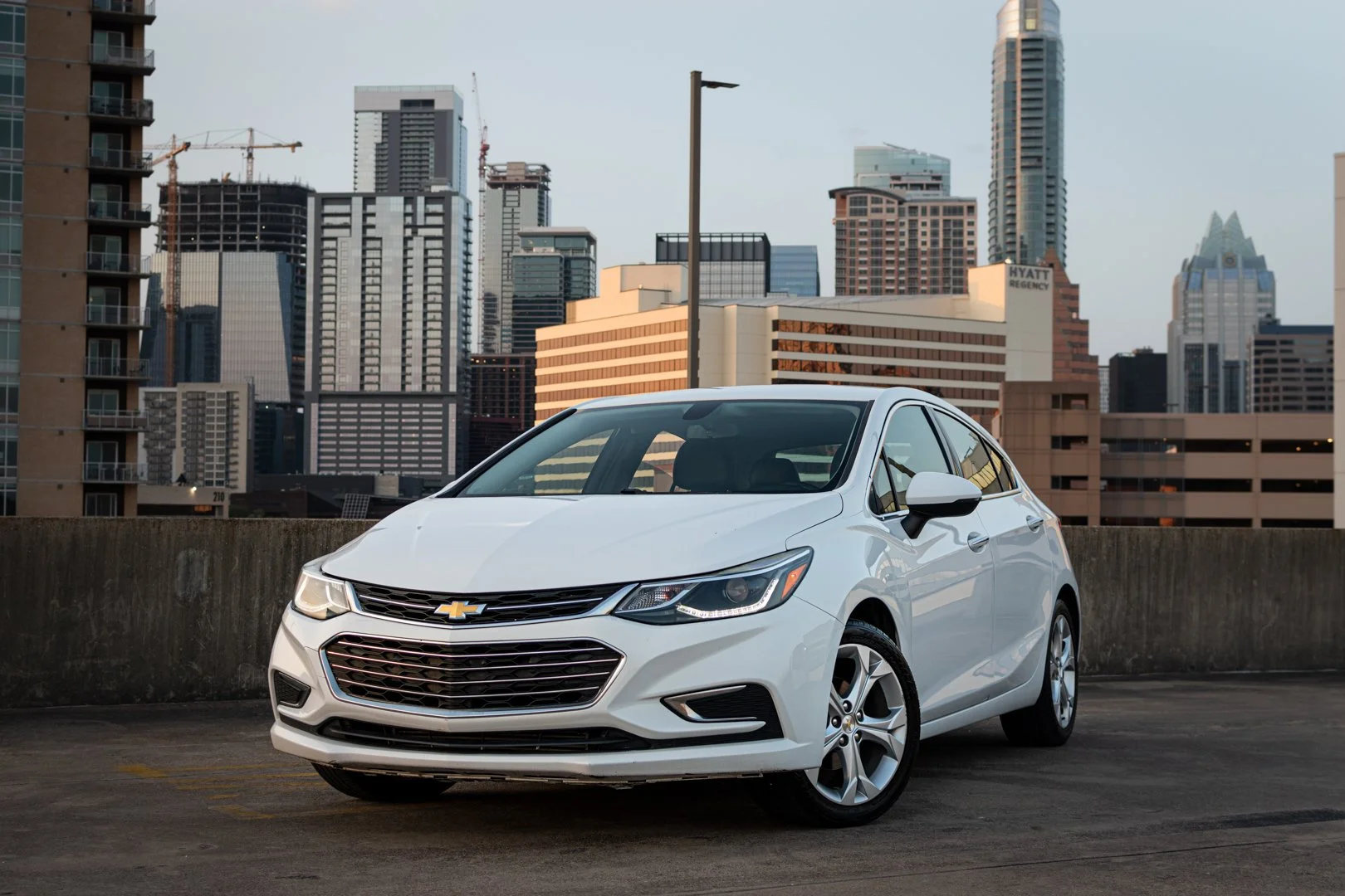 A white Chevrolet car parked on the rooftop parking lot of a city with many tall skyscrapers in the background.