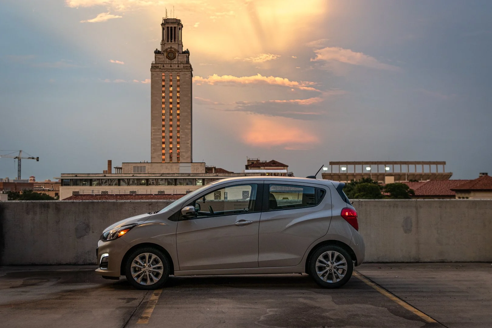 A grey compact car parked on the rooftop parking lot at sunset with a tall clock tower and city skyline in the background.
