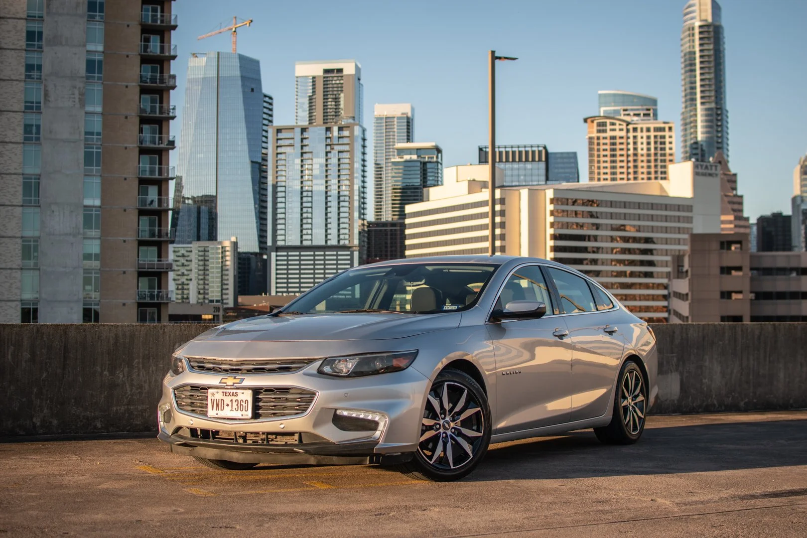 Silver Chevrolet sedan parked on a rooftop parking lot with a city skyline in the background during sunset.