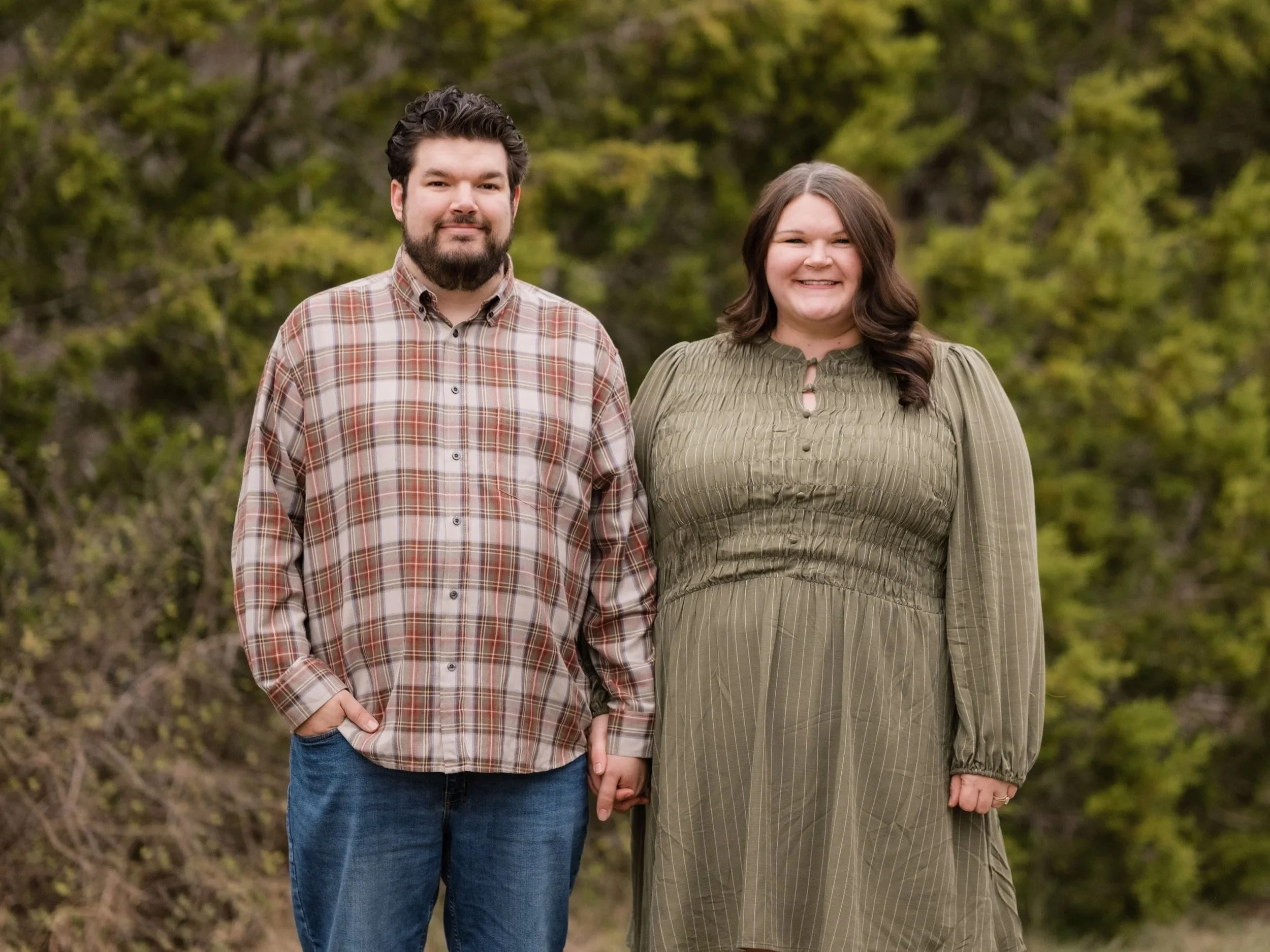 A man and woman standing together outdoors, holding hands, with green trees in the background.