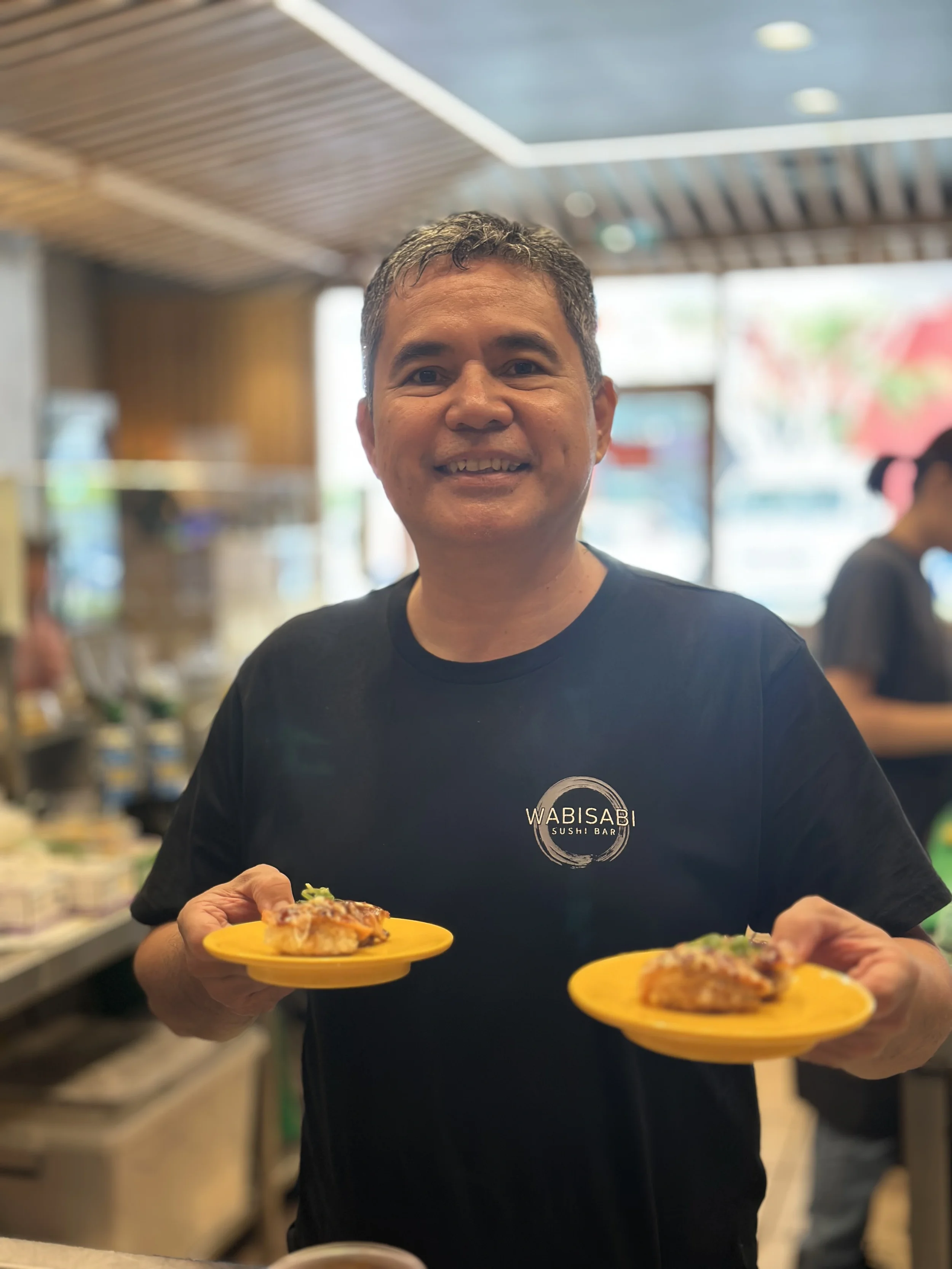 Smiling man holding two yellow plates with sushi at a sushi bar restaurant.