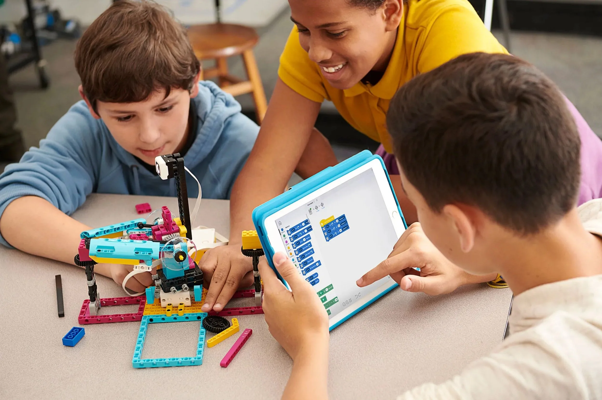 Three children and a young woman assembling a LEGO robotics project on a table, with a tablet displaying programming code in the foreground.