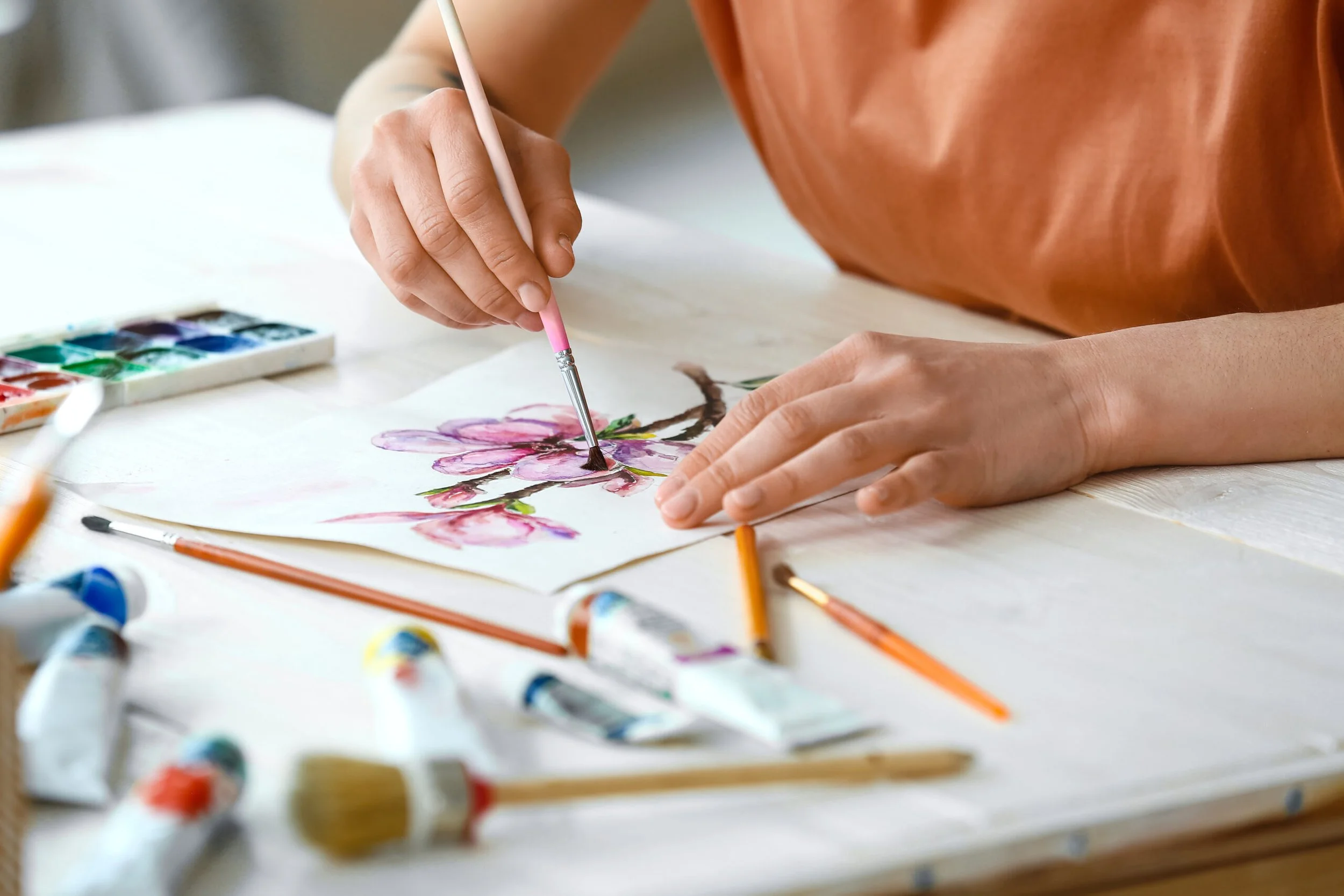 Person painting pink cherry blossoms on paper with watercolor paints and brushes.