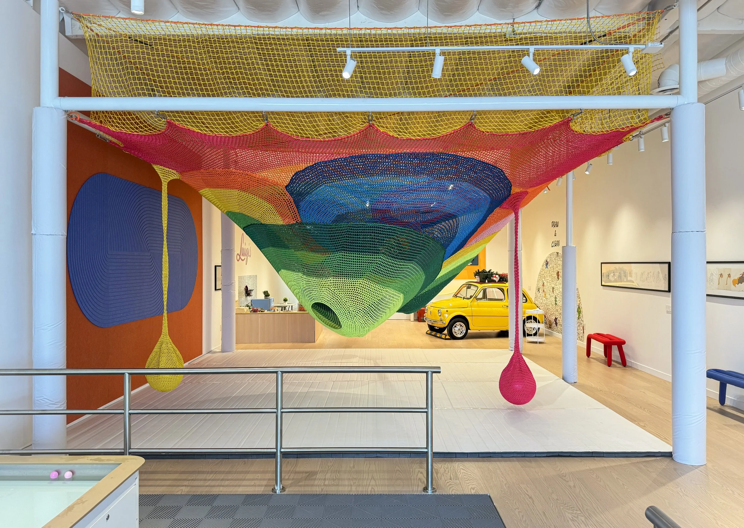 Colorful woven art installation with a rainbow gradient pattern, suspended from a ceiling in an indoor space with various artworks, a vintage yellow car, a pool table, and seating visible in the background.