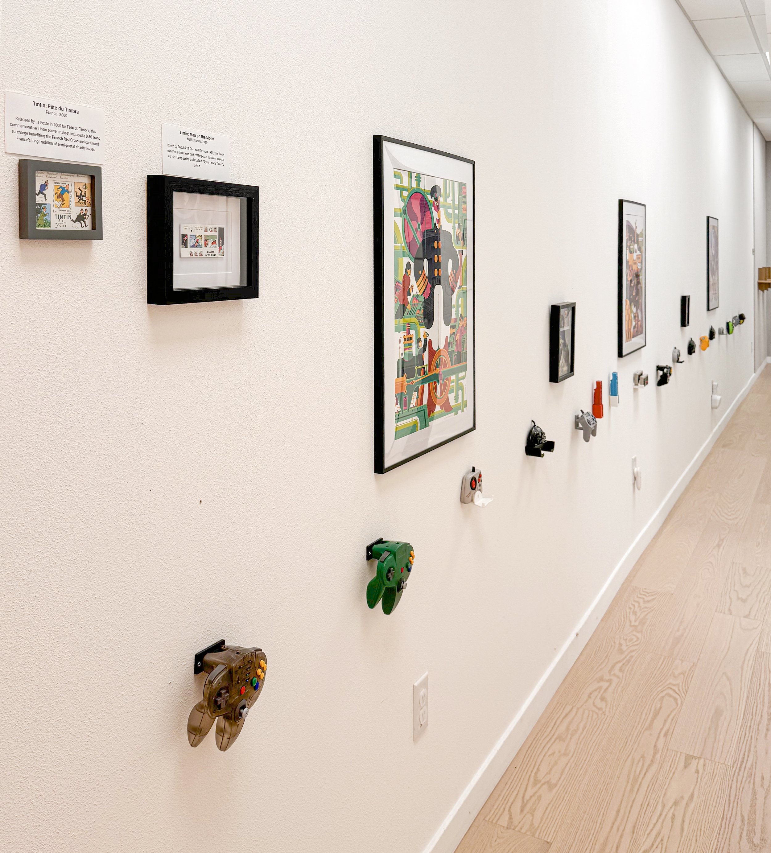 Art gallery wall display featuring framed artwork, with 8-handheld gaming controllers mounted below, arranged in a line along a white wall.