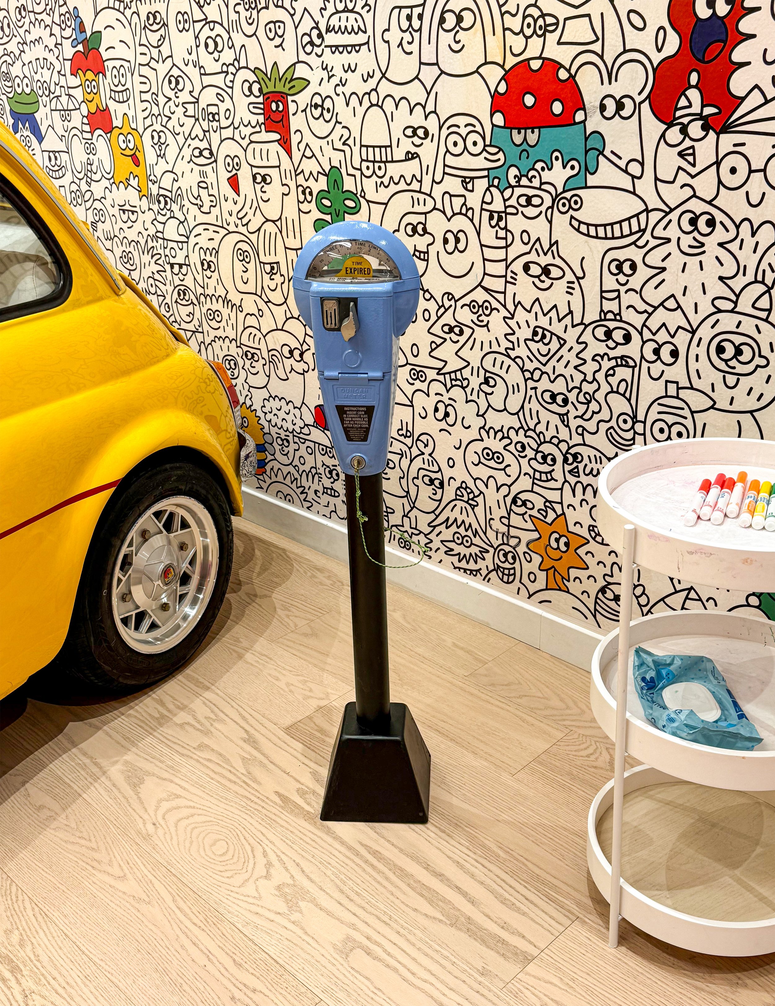 A colorful wall mural featuring numerous cartoon characters and faces in black and white, with some characters colored in red, yellow, blue, and green. A yellow car is partially visible on the left, and a blue parking meter with a sign indicating 'Ex