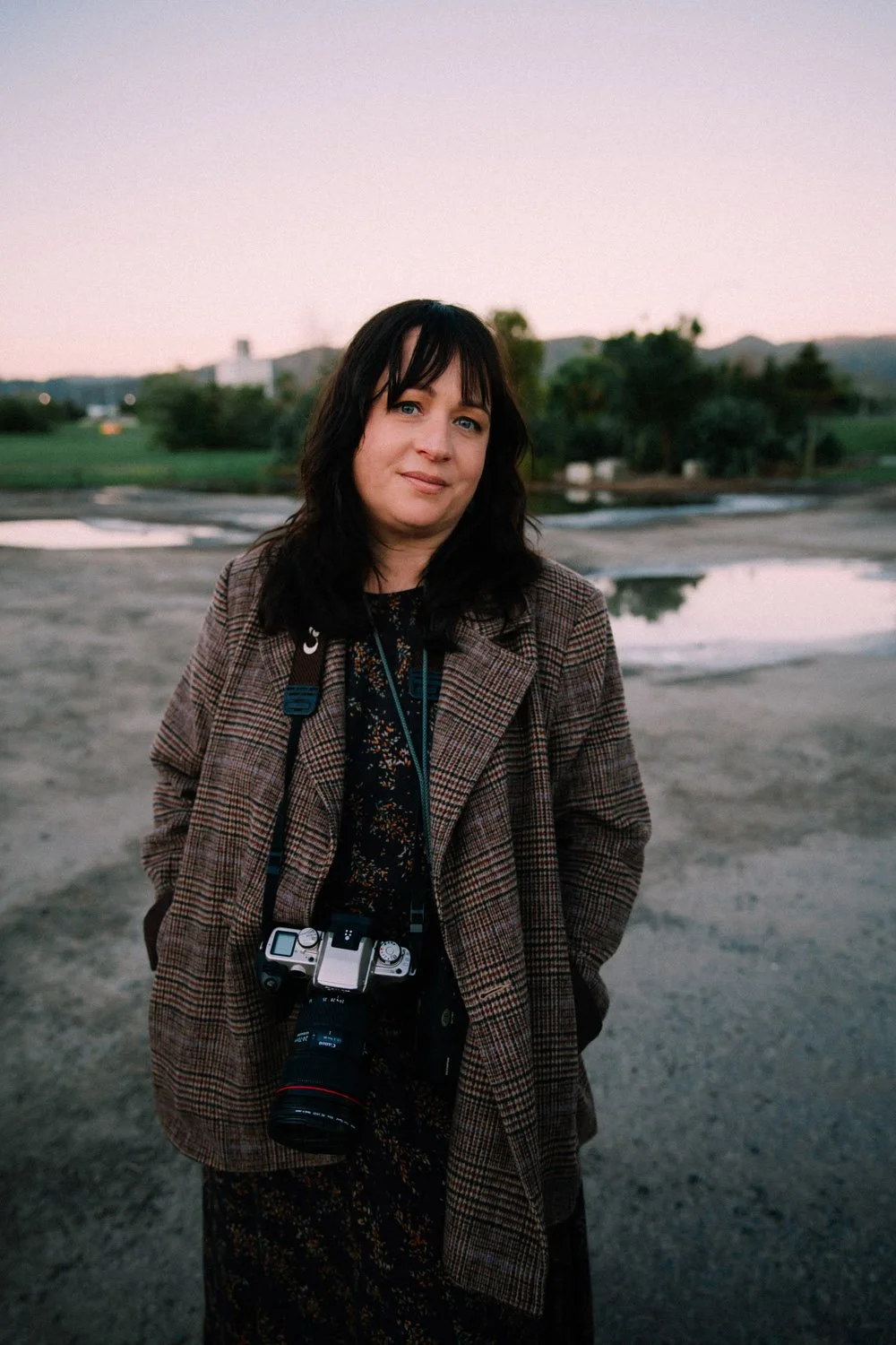 Sarah McEvoy wearing a plaid coat and a camera around her neck outdoors during dusk, with a landscape of trees, hills, and puddles in the background.