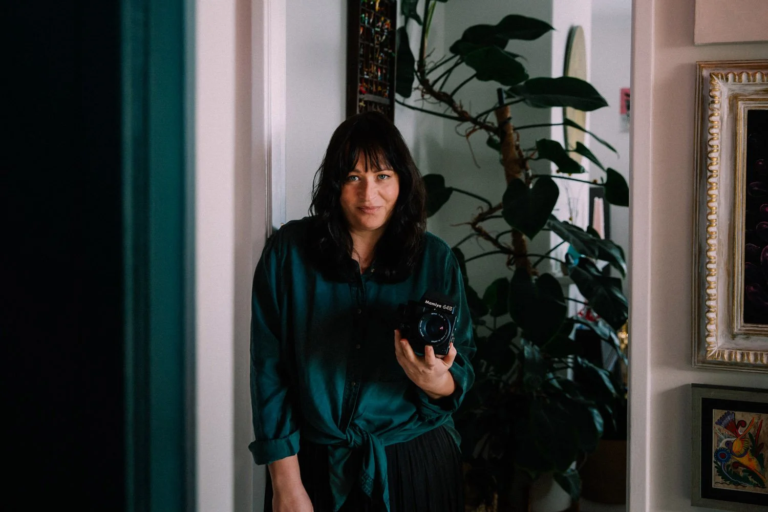 Sarah McEvoy, wearing a teal shirt, with a camera in her hand. In the background, there is a large plant and framed artwork on the wall.