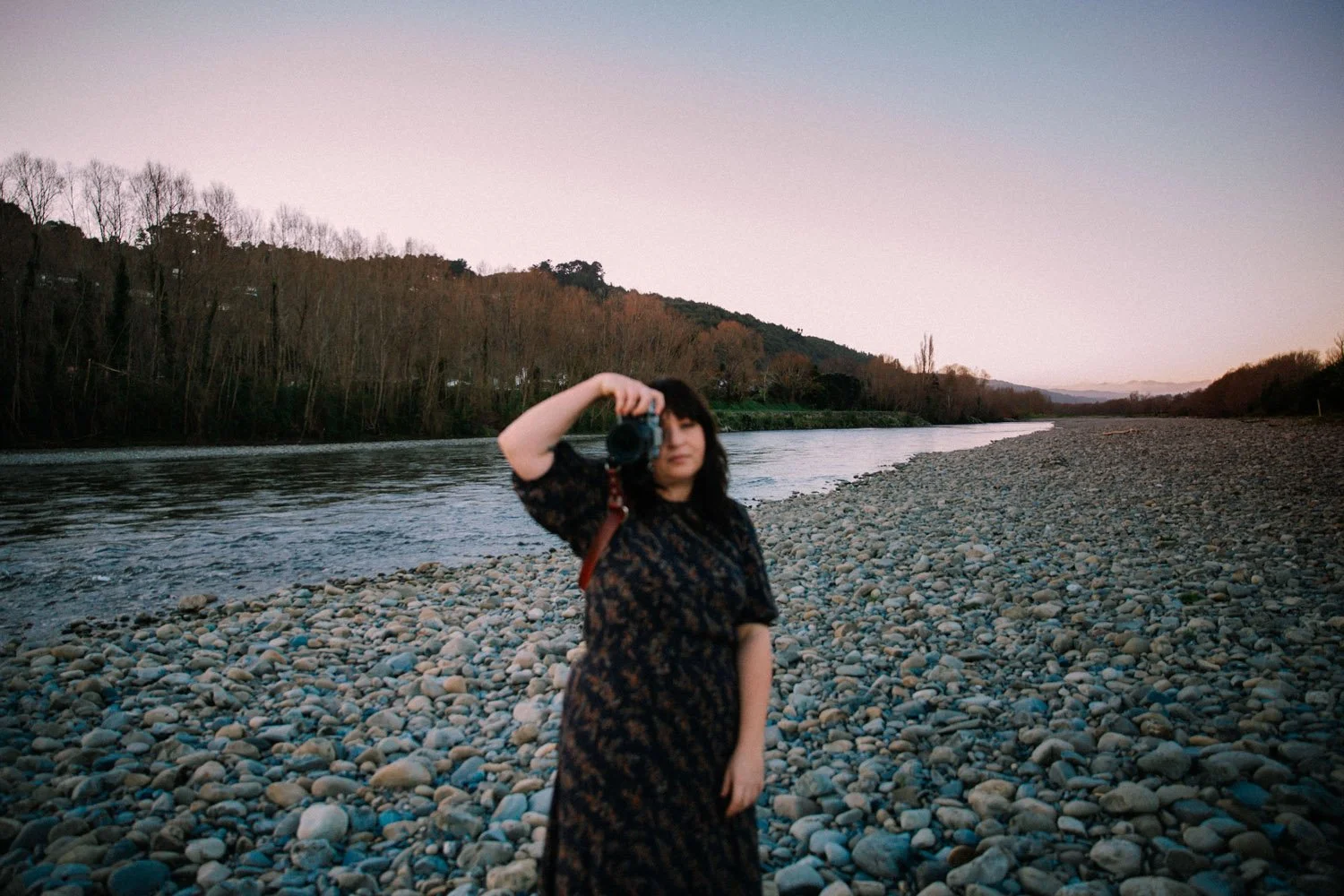 Sarah McEvoy in a black dress taking a photo with a camera on the Hutt river at sunset.