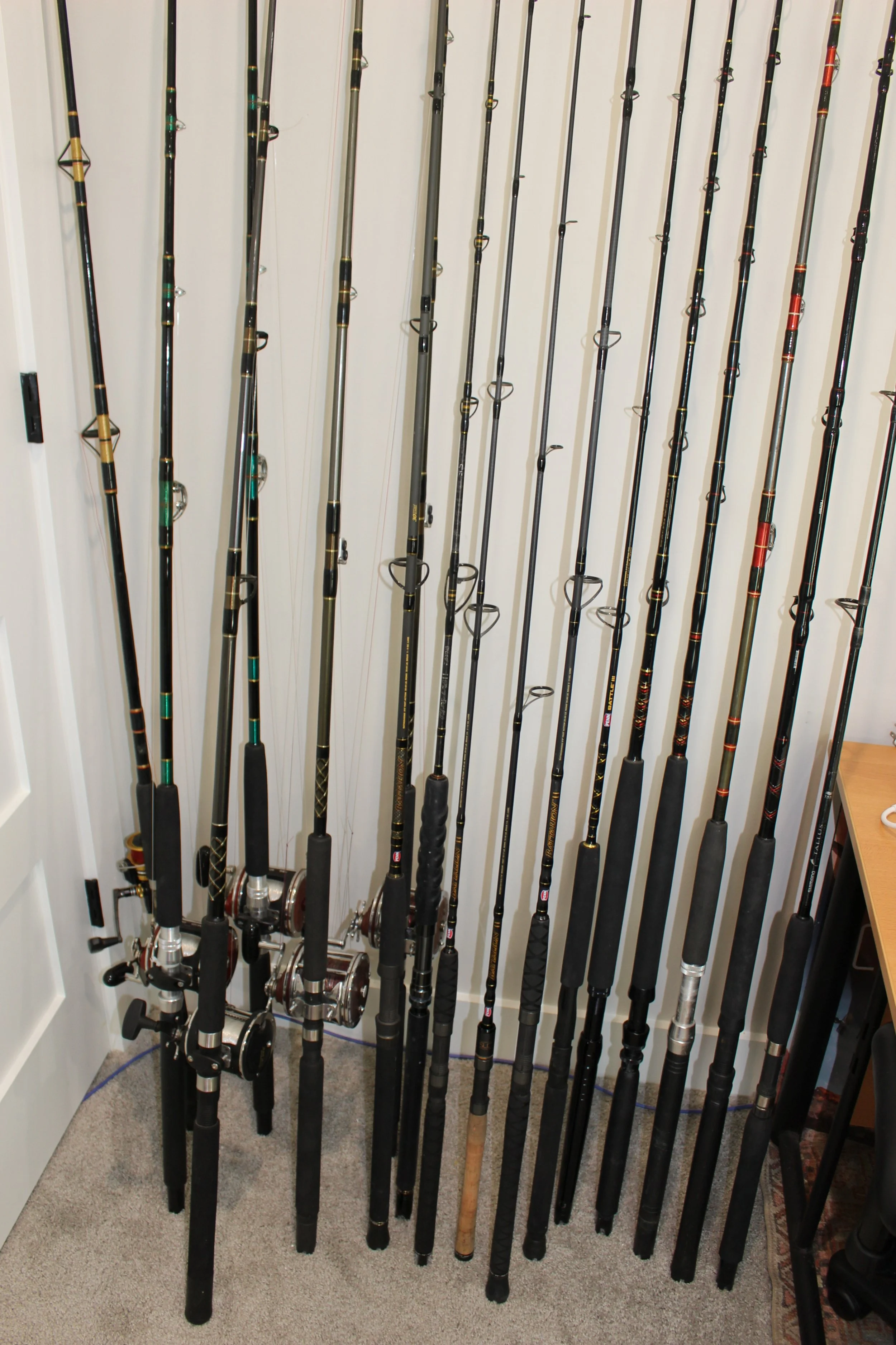 Several fishing rods lined up against a white wall in a room, with some rods resting on a small table and on the floor.