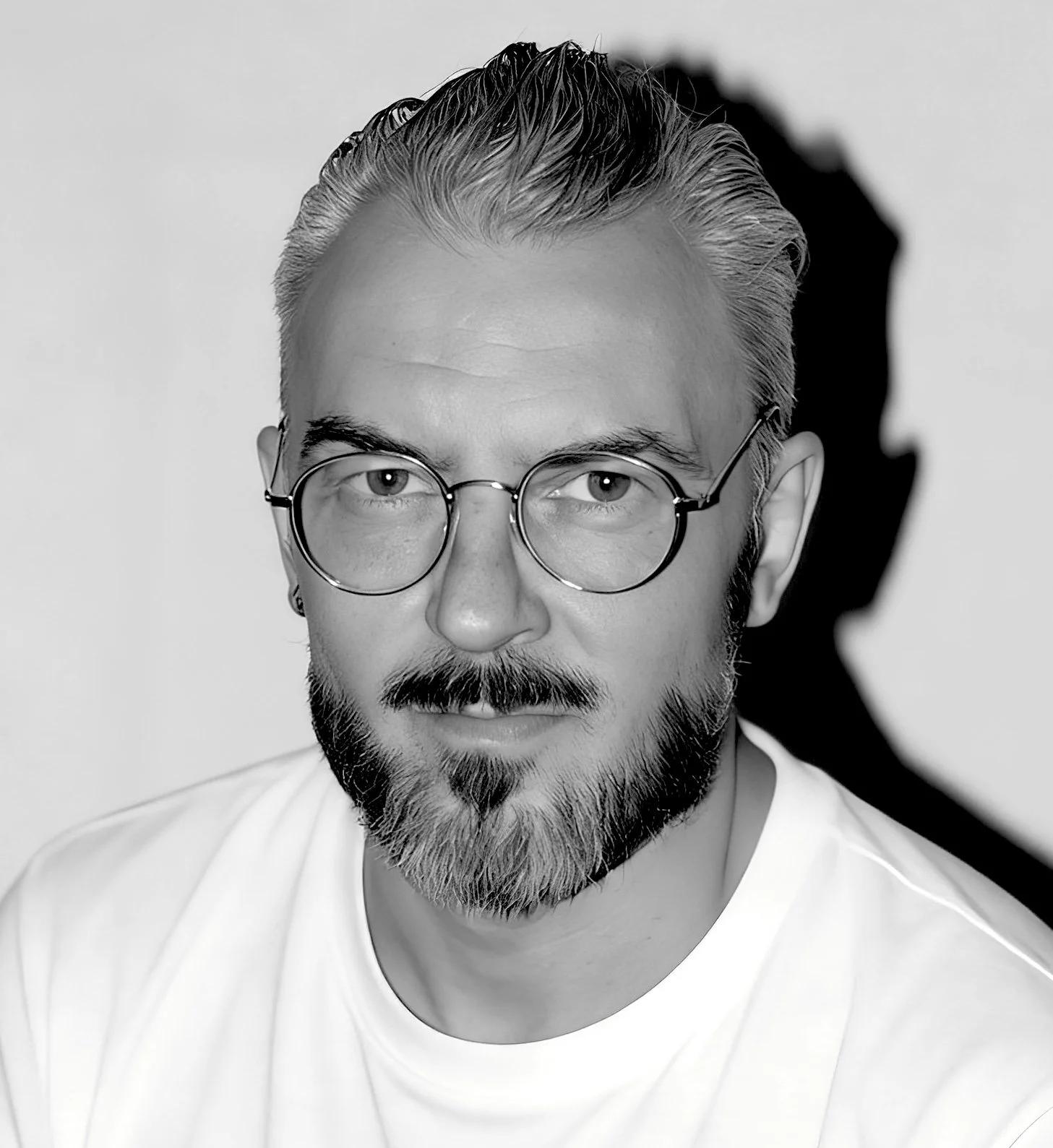 Black and white portrait of a man with glasses and a beard, wearing a white shirt, looking directly at the camera with a serious expression.