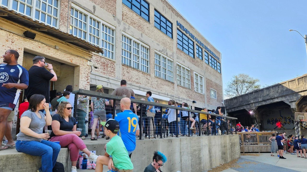 An outdoor view of a lively gathering at the Ravine. Numerous people are hanging out on an elevated patio, while others sit on the concrete retaining wall.