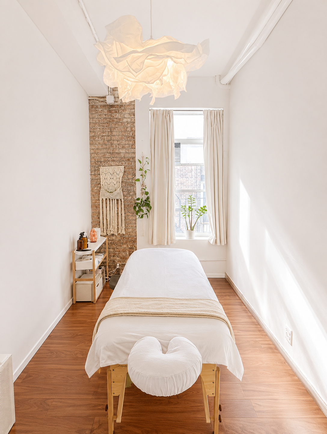 A wellness or massage room with a massage table covered in white sheets, a large window with cream curtains, plants near the window, a small side table with essential oils, a hanging macrame wall decoration, in press massage union square nyc