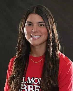 Young woman with long dark hair smiling, wearing a red shirt with white text, against a dark background.