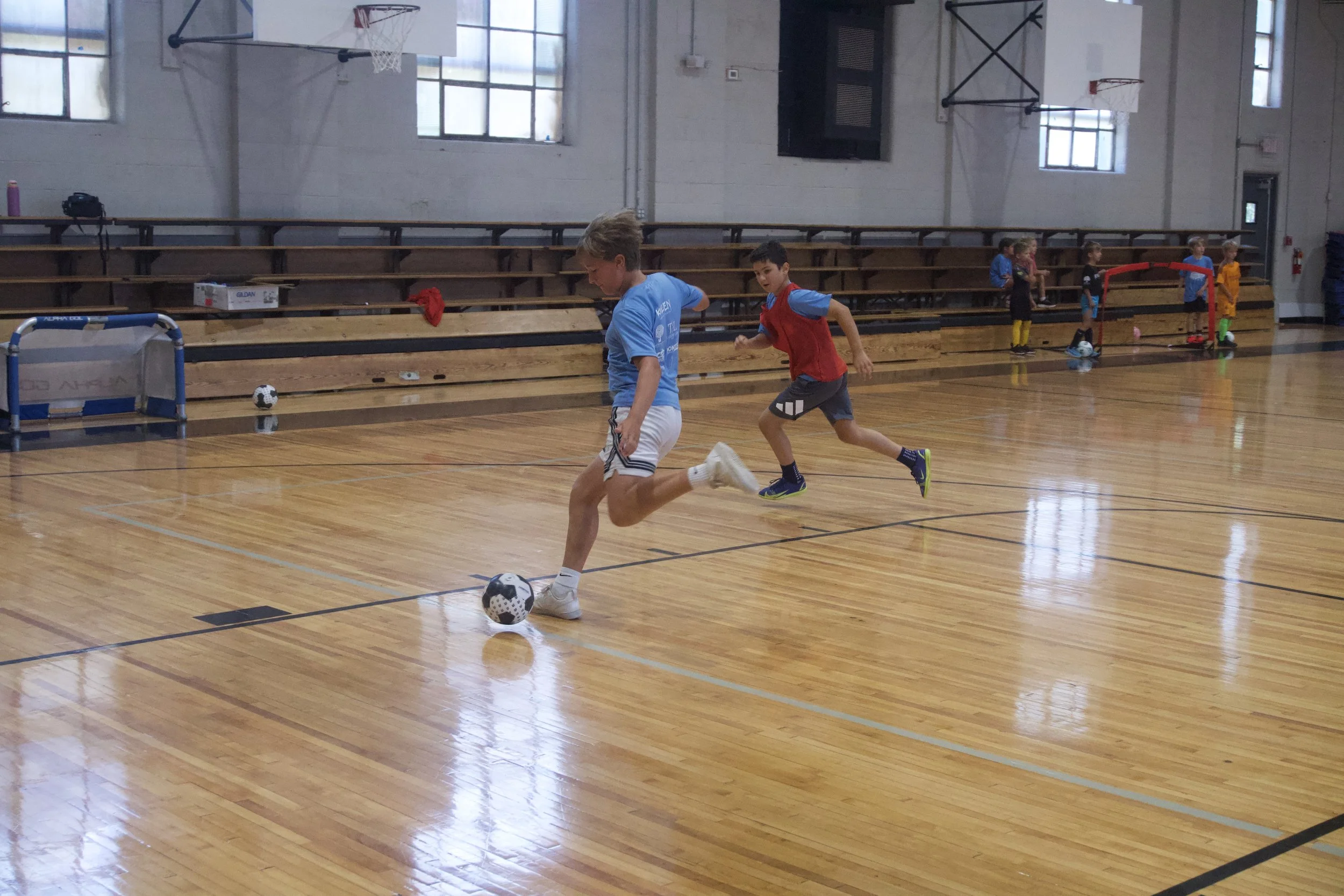 Children playing indoor soccer in a gymnasium, with one child kicking a soccer ball while others watch from the background.