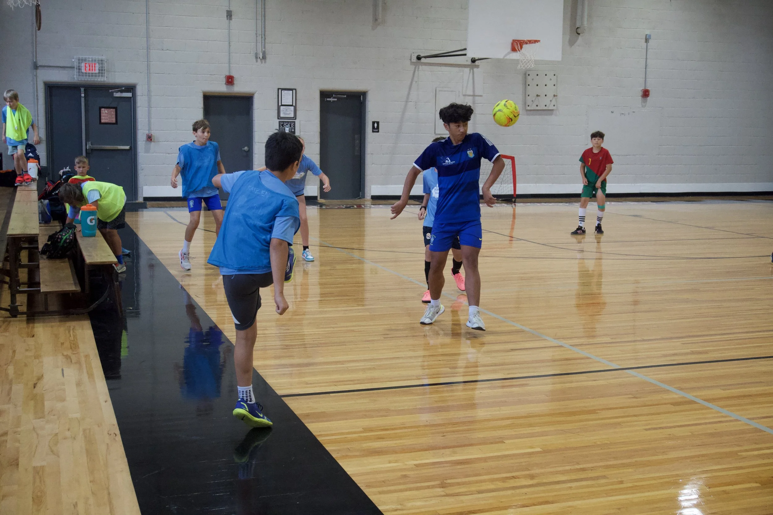 Children playing indoor soccer in a gymnasium with wooden floors and a basketball hoop in the background.