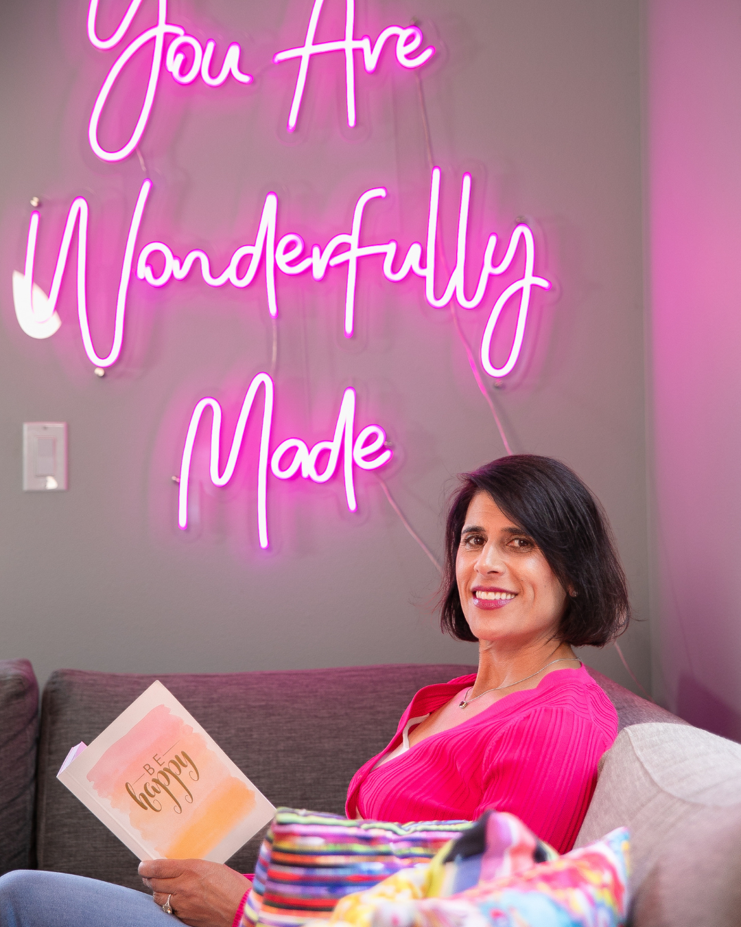 Heidi Carlson, your coach, sitting on a gray couch, holding a book that says 'Be Happy'. Behind her, a neon pink sign on the wall reads 'You Are Wonderfully Made'.