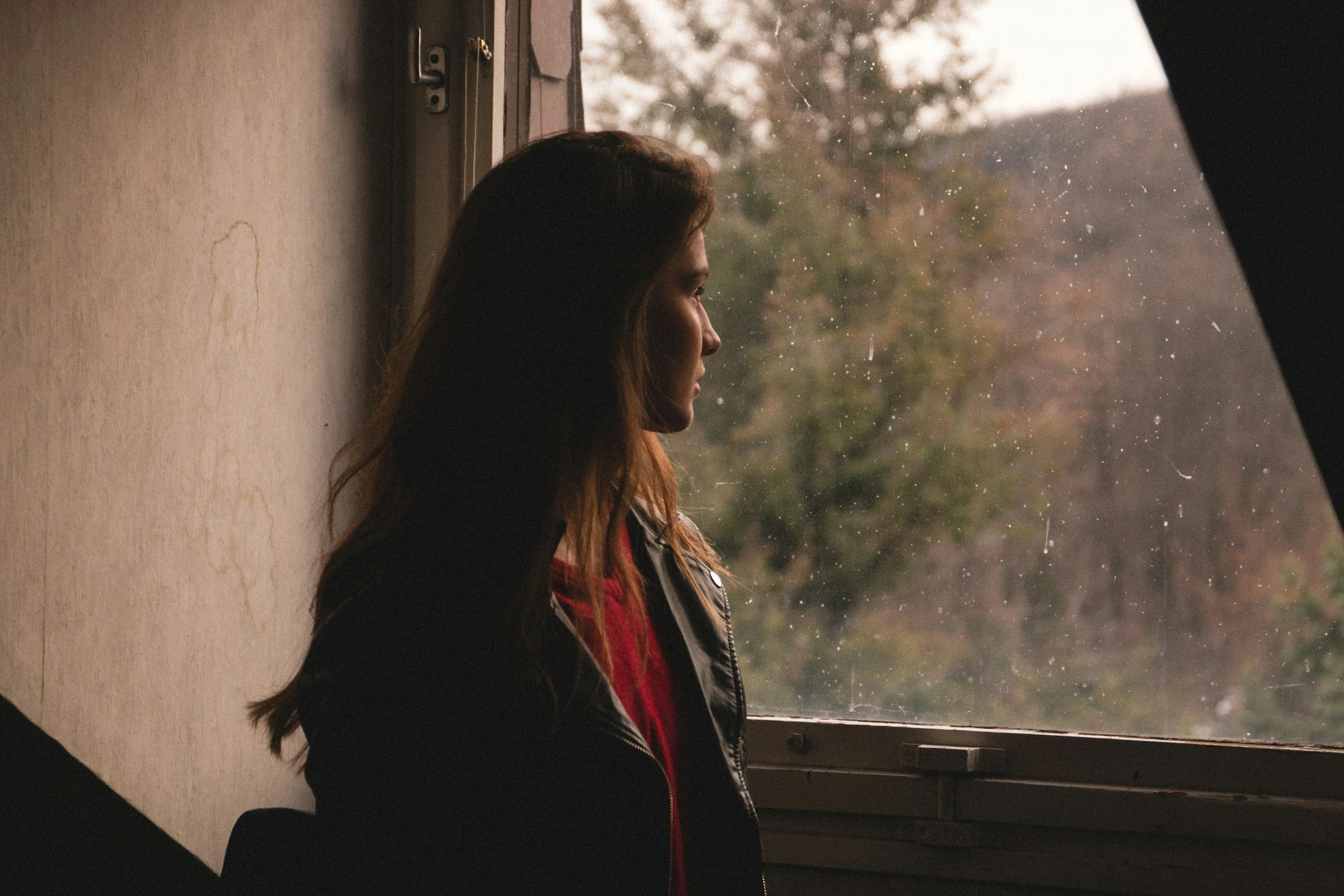 A woman with long hair and wearing a black jacket sitting by a window, looking outside on a rainy day.