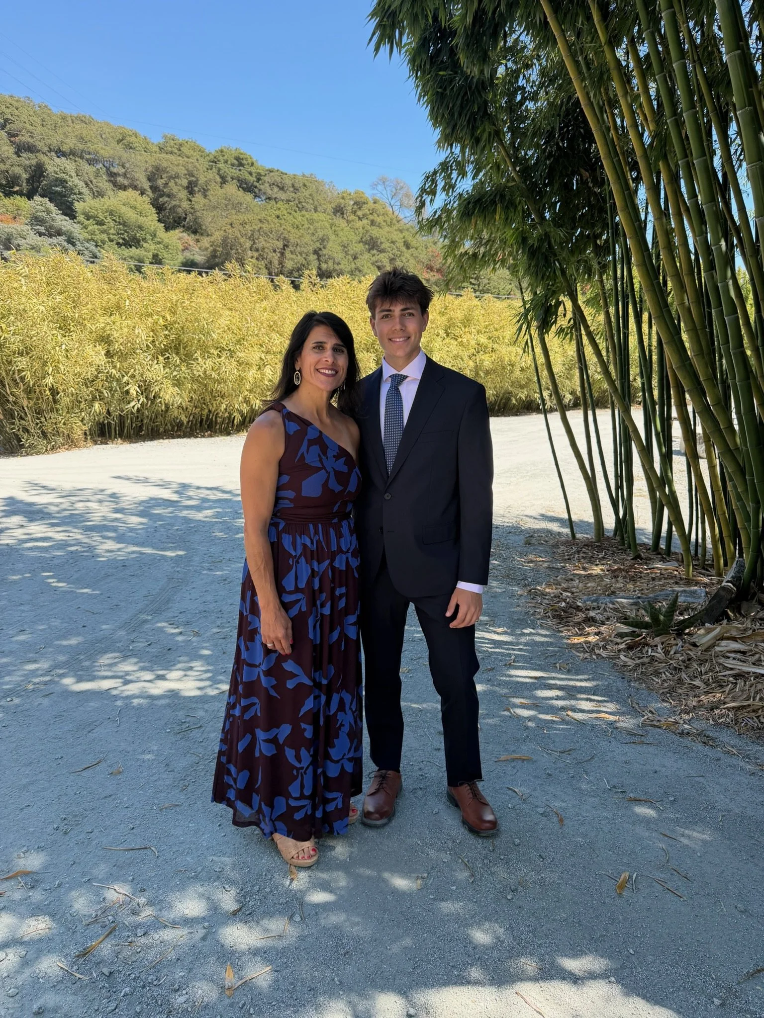 A woman and a man dressed in formal attire standing outdoors on a sunny day with yellow foliage, trees, and a blue sky in the background.