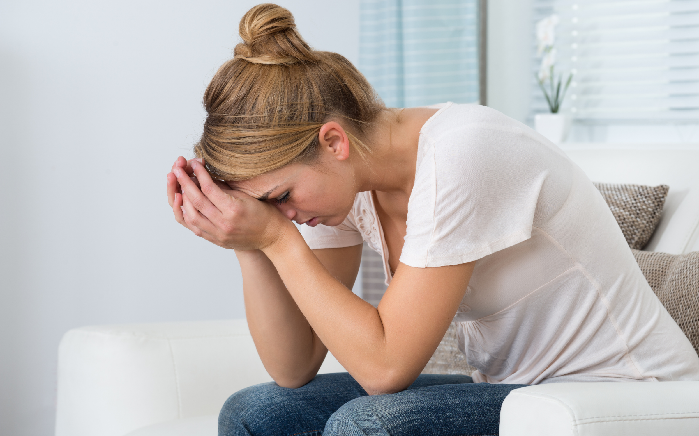 A woman with blonde hair tied in a bun, sitting on a white couch with her head bowed and hands covering her face, appears distressed or upset in a bright room with a white wall, a window with blinds, and a potted plant in the background.
