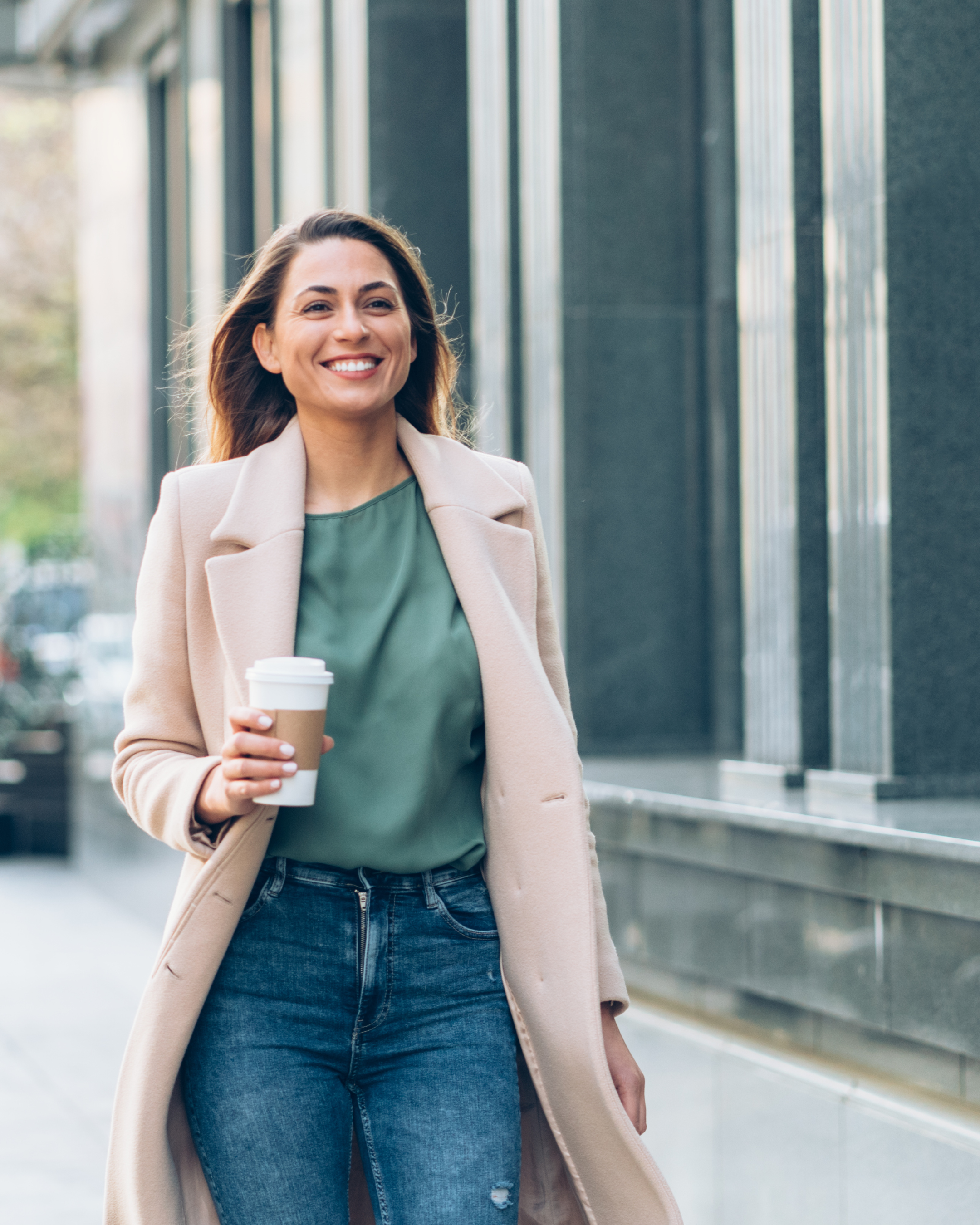 A woman smiling and holding a coffee cup while walking outdoors feeling confident, relieved, and free.