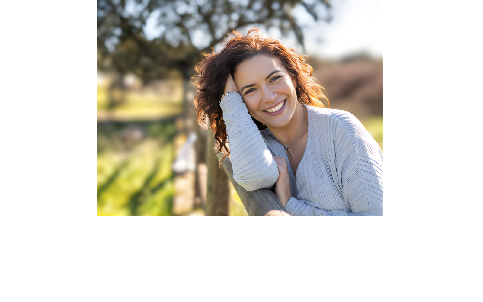 A woman with curly brown hair smiling outdoors, leaning her head on her hand, with a blurred green park background.