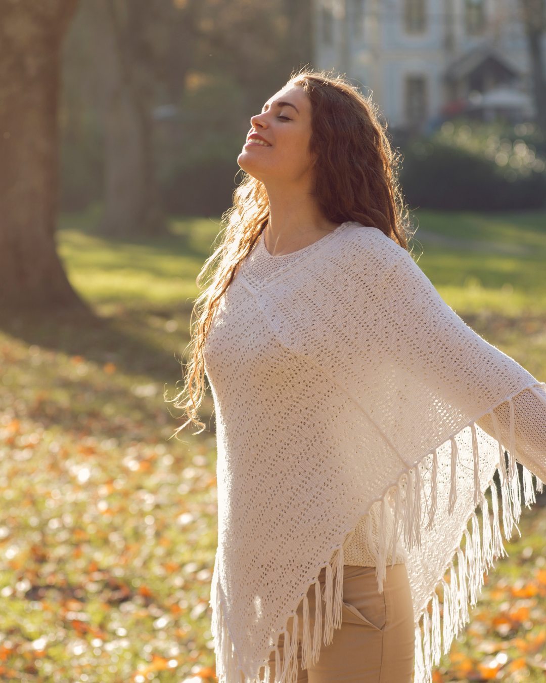 Woman with long curly hair enjoying sunlight outdoors in a park, wearing a cream crochet poncho with fringe details and beige pants.