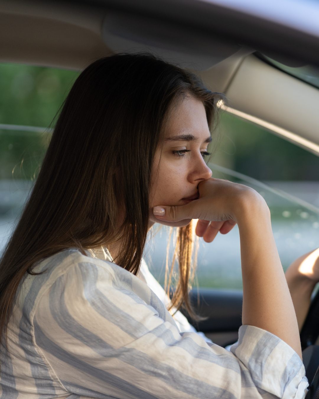 A woman with long brown hair resting her chin on her hand inside a car, appearing contemplative or bored.