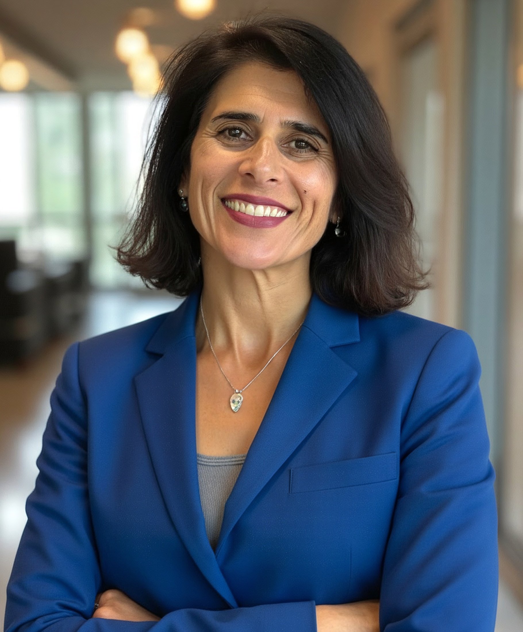 A smiling woman with dark hair wearing a blue blazer and jewelry in a professional indoor setting.