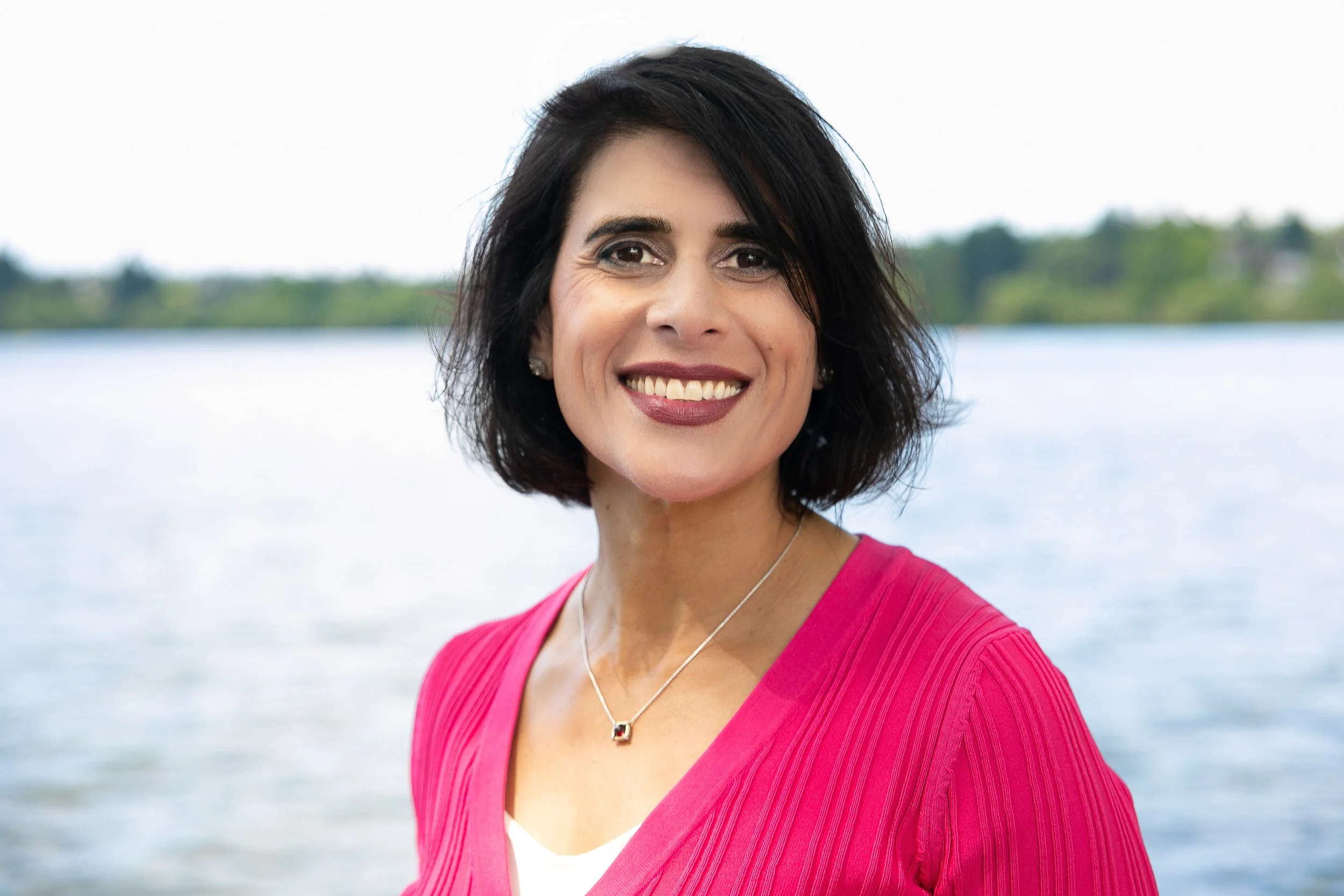 A woman with short dark hair and a pink top standing outdoors near a body of water, smiling at the camera.