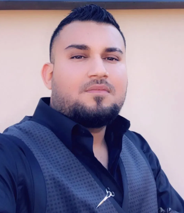 A man with dark hair and facial hair taking a selfie outdoors, wearing a blue shirt and a backpack.