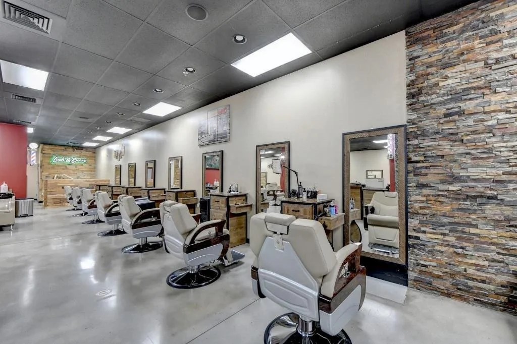 An interior view of a modern barbershop or salon with multiple styling chairs, mirrors, and wooden workstations along a white wall. The space has polished concrete floors, a stone accent wall, and ceiling lights.