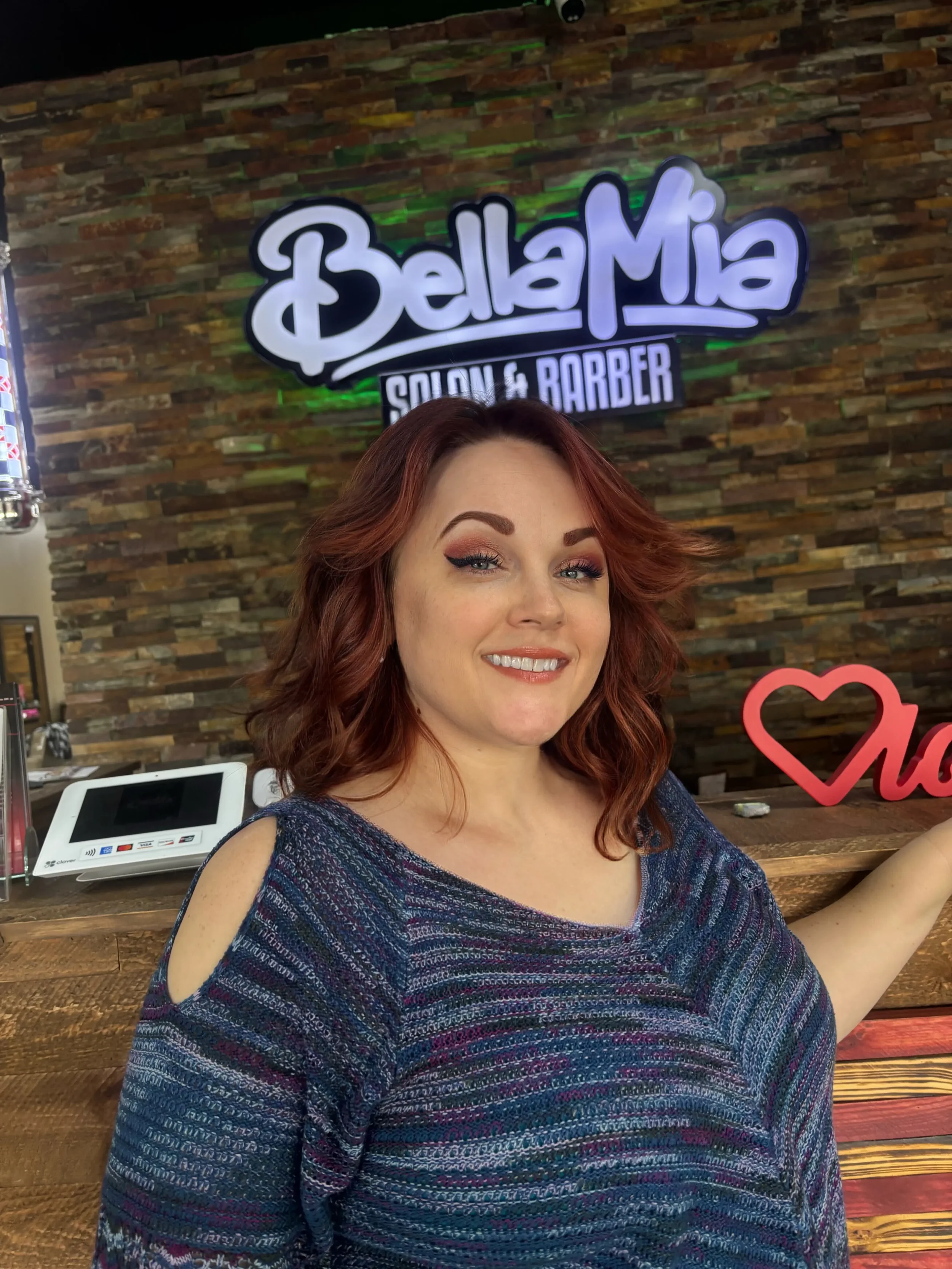 A woman with red hair and makeup, smiling at the camera, in front of a wall with a sign that reads 'Bella Mia Salon & Barber' and a decorative red heart, with a point-of-sale system visible on the counter.