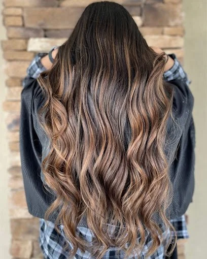 Back view of a woman with long, wavy, ombre hair transitioning from dark brown at the roots to light brown at the ends, standing in front of a brick wall.