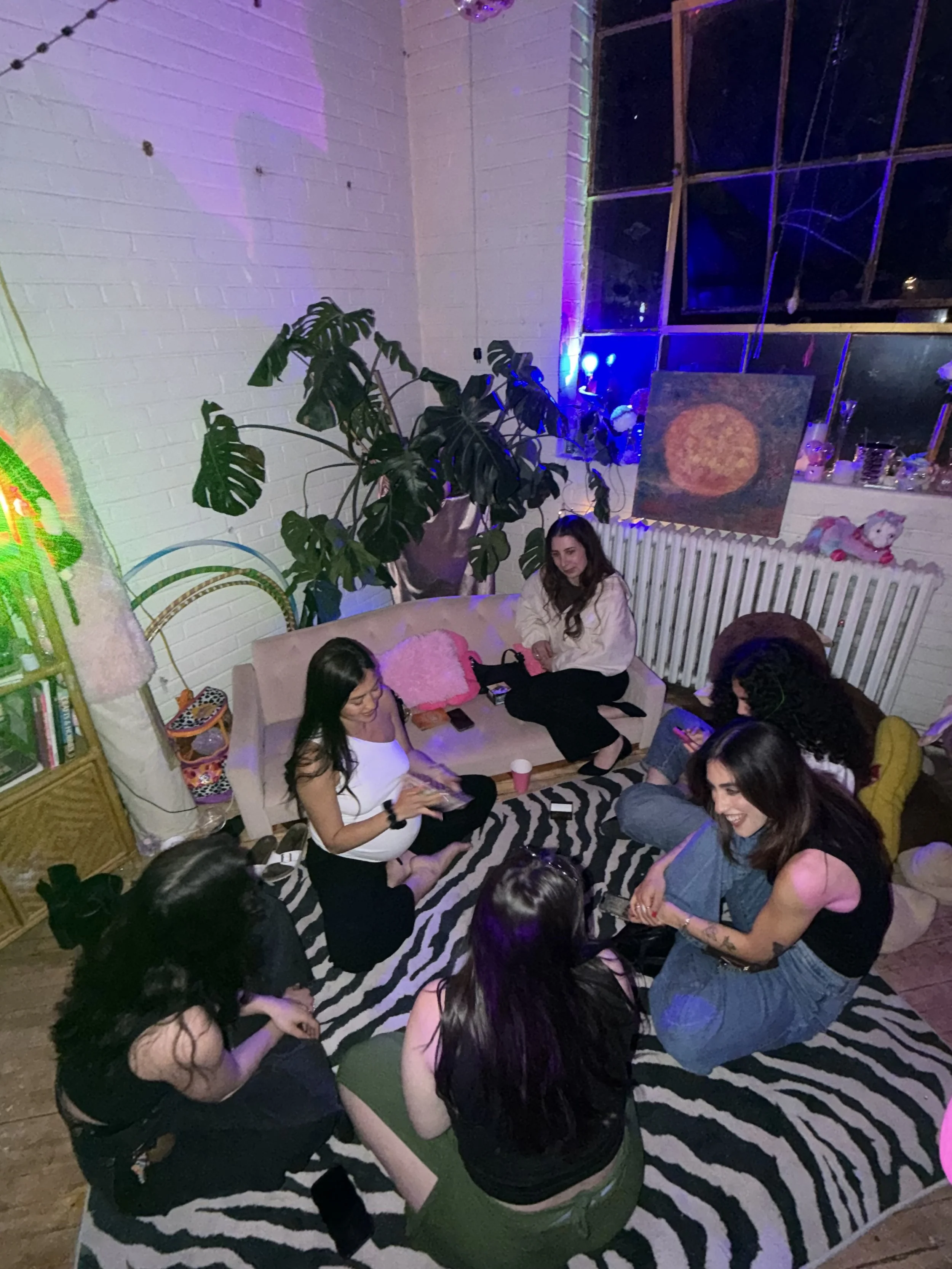Group of women sitting on a zebra print rug in a cozy room, chatting and smiling. One pregnant woman in the center is in a white top and black pants, while others wear variety of casual clothing. The room has a large window, a plant, wall art, a radiator, and shelves with decor. The lighting is dim and colorful.