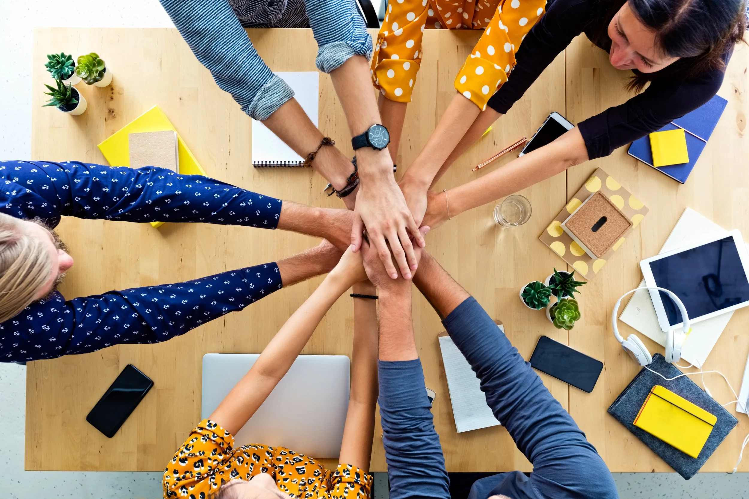People in a meeting place their hands together in the center of a wooden table, surrounded by laptops, tablets, smartphones, notebooks, small potted plants, and office supplies.