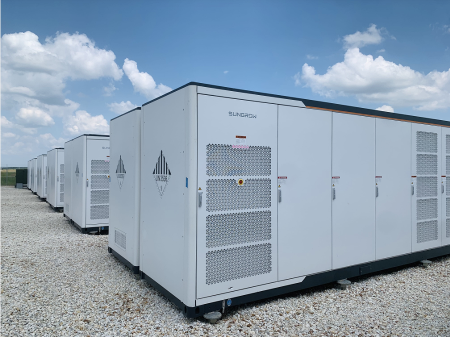Typical BESS unit installed within a BESS site. The white metal container is approximately 6 metres wide, 3 metres tall and 2.5 metres deep. Gravel pathways surround the unit, with four additional BESS units arranged in rows behind it.