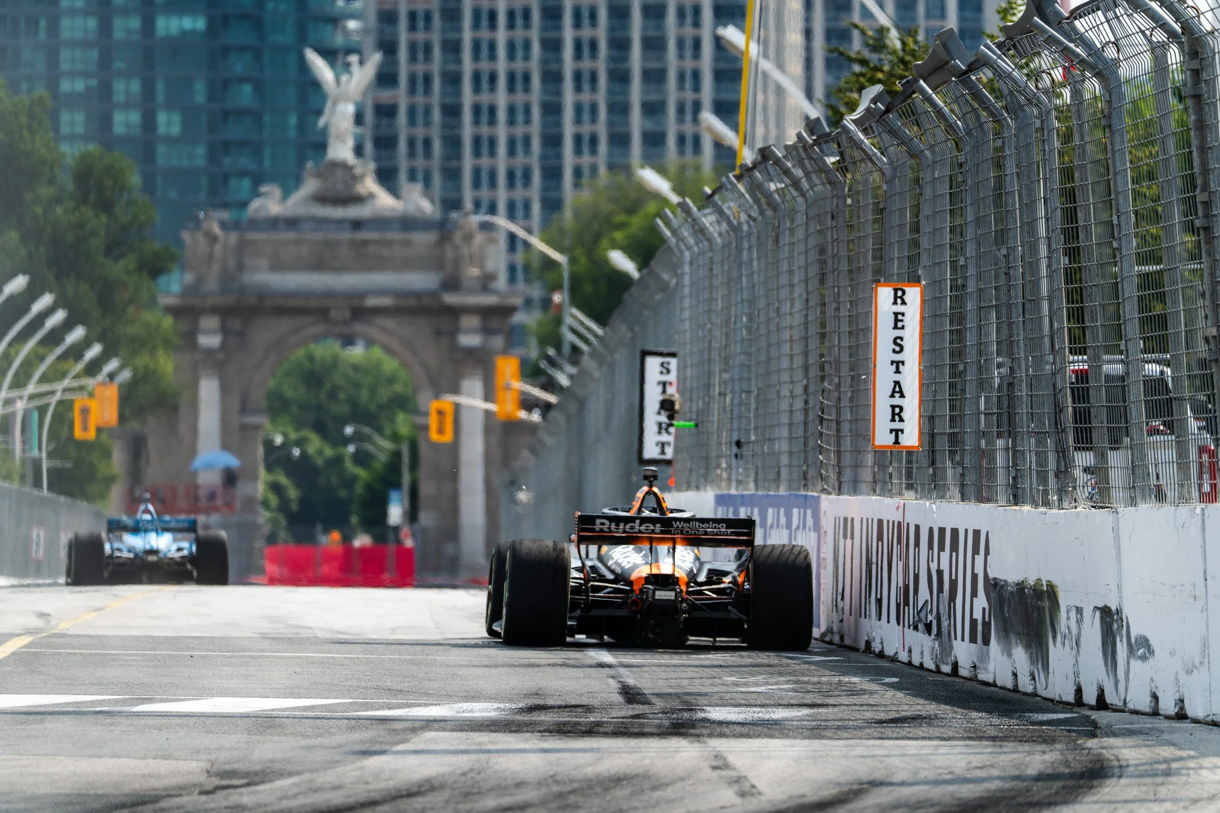 A racing car on a street circuit near a city arch landmark, with barriers and safety fencing. The car is finishing a race on a sunny day with trees and buildings in the background.