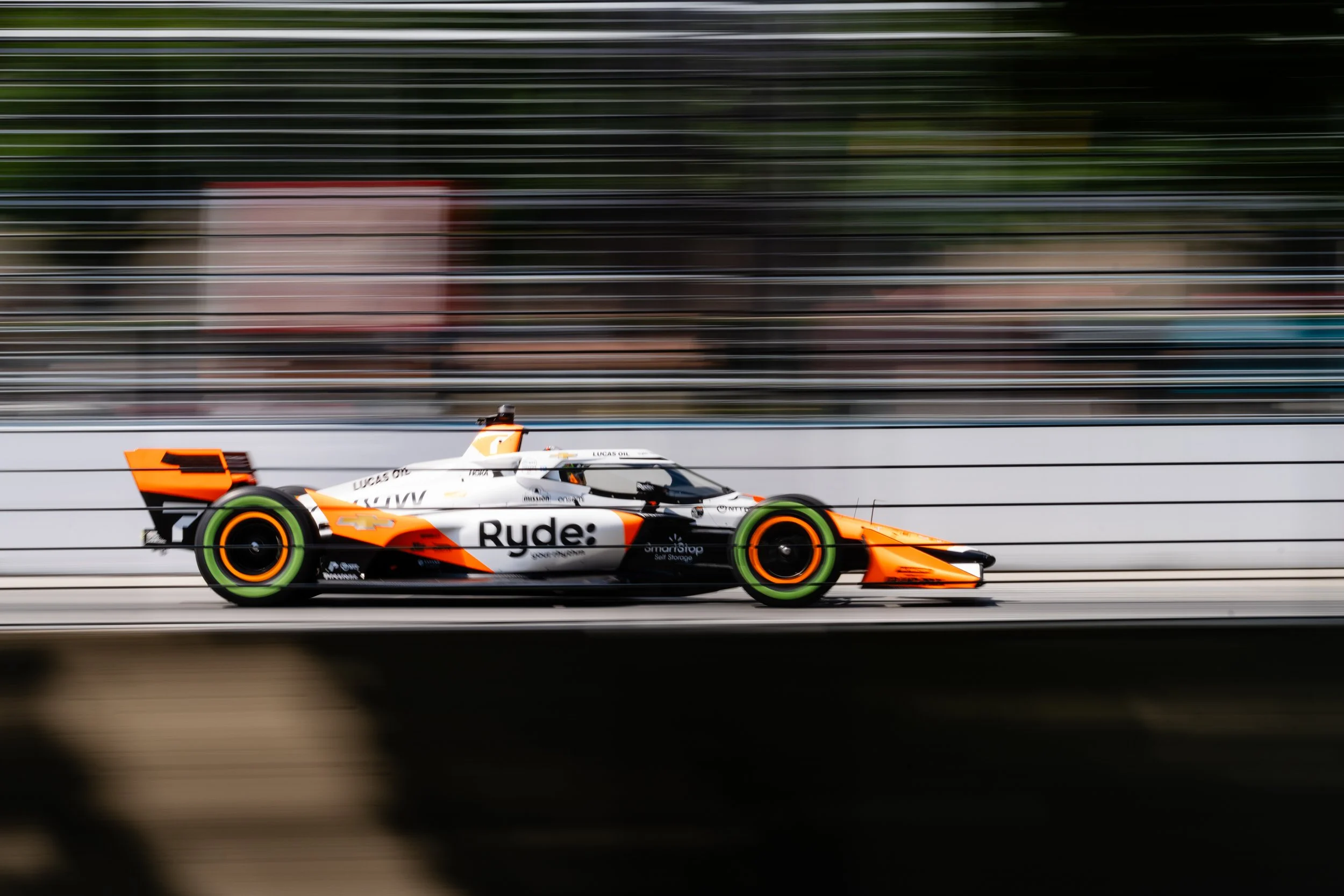 A race car moves quickly on a track, with motion blur background, painted in white, orange, and black, displaying sponsor logos including 'Ryde:' and Chevrolet, with neon green rims.