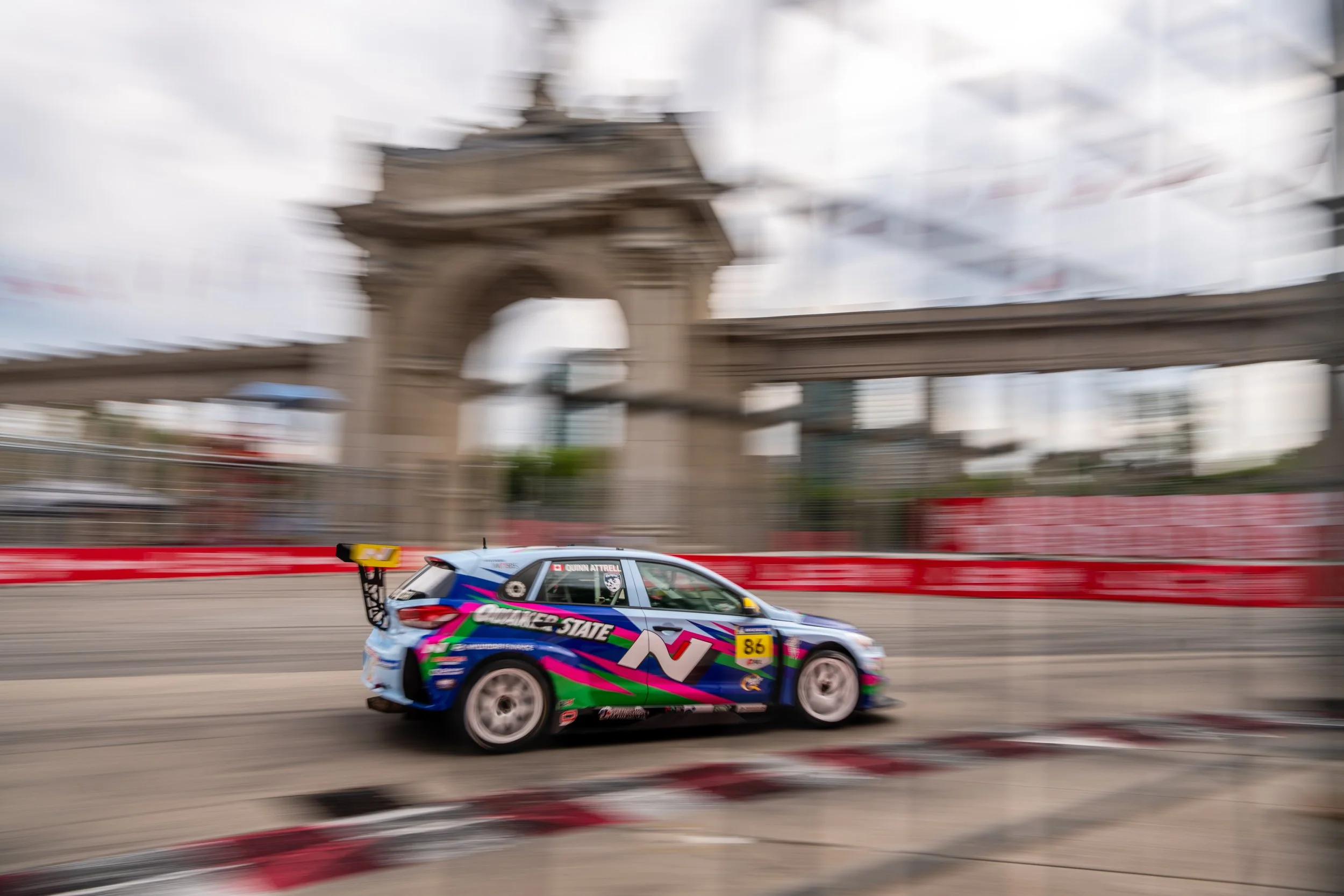 Veloster race car racing on the streets of toronto during the Indycar race weekend, with a blurred background of city architecture and a cloudy sky.
