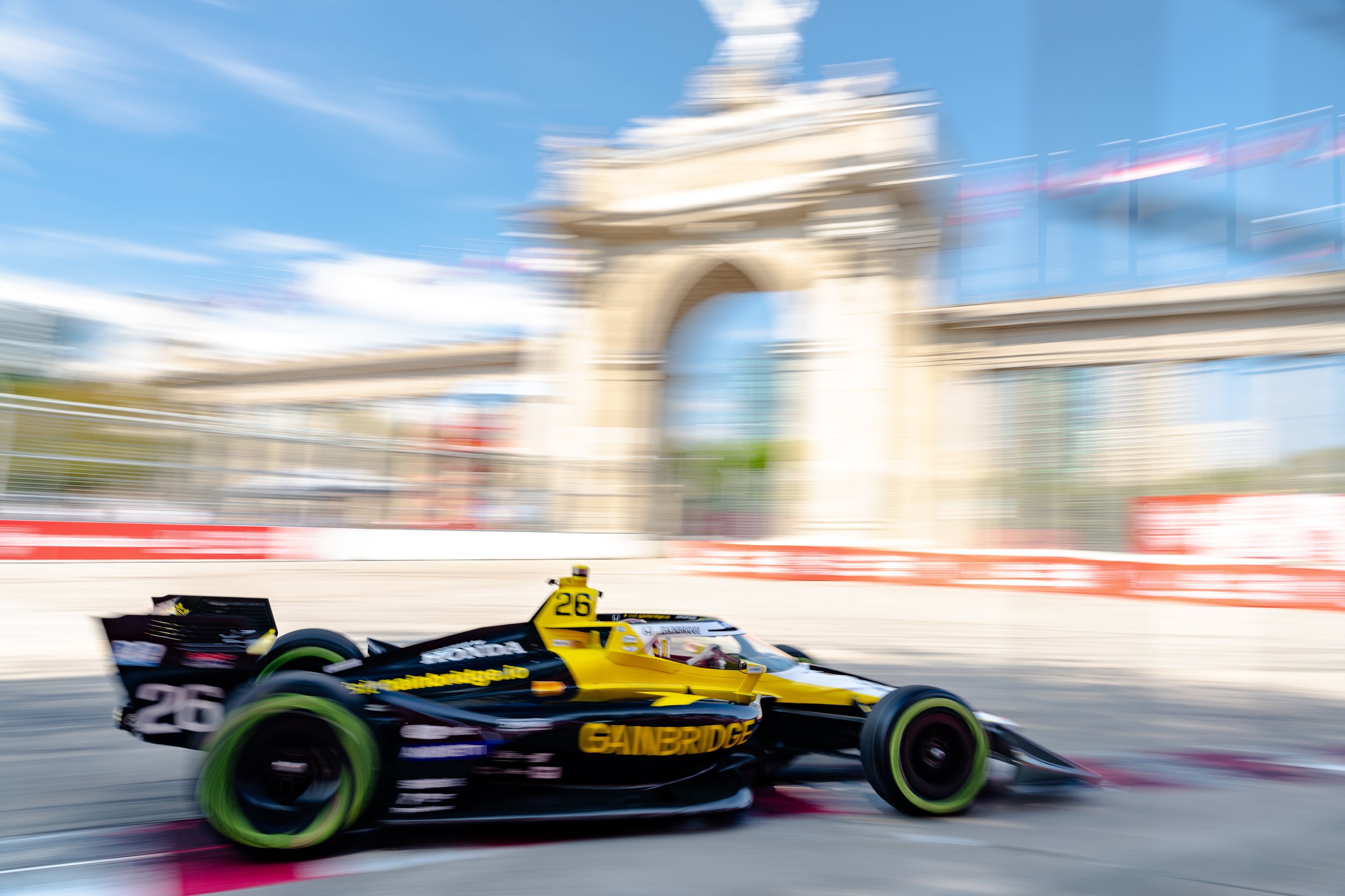 Blurred image of a yellow and black IndyCar race car speeding on a track with a large arch and blue sky in the background.