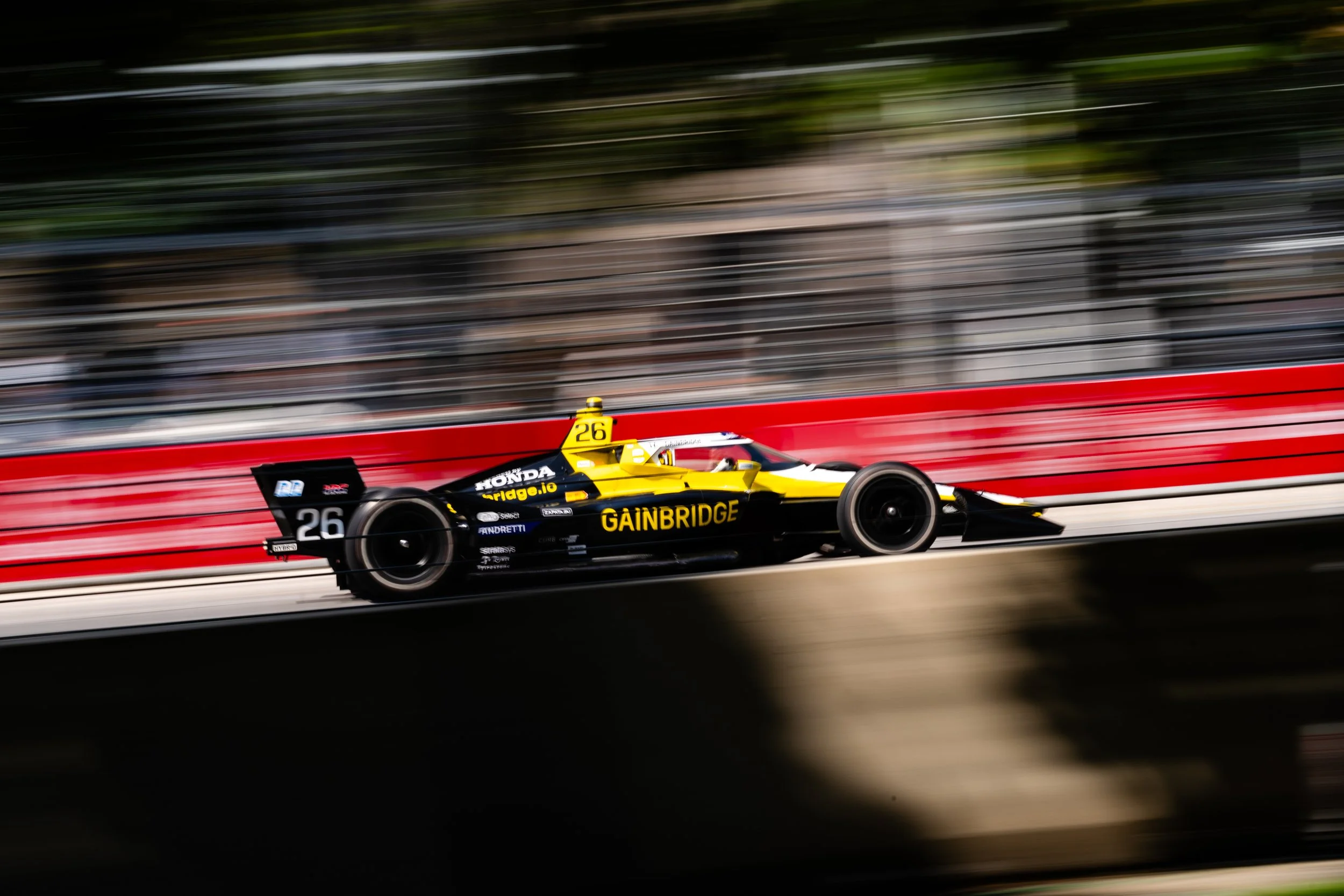 A yellow and black Formula racing car with the number 26, sponsored by Gainbridge, rushing along a racetrack with a red and white barrier and a blurred background.