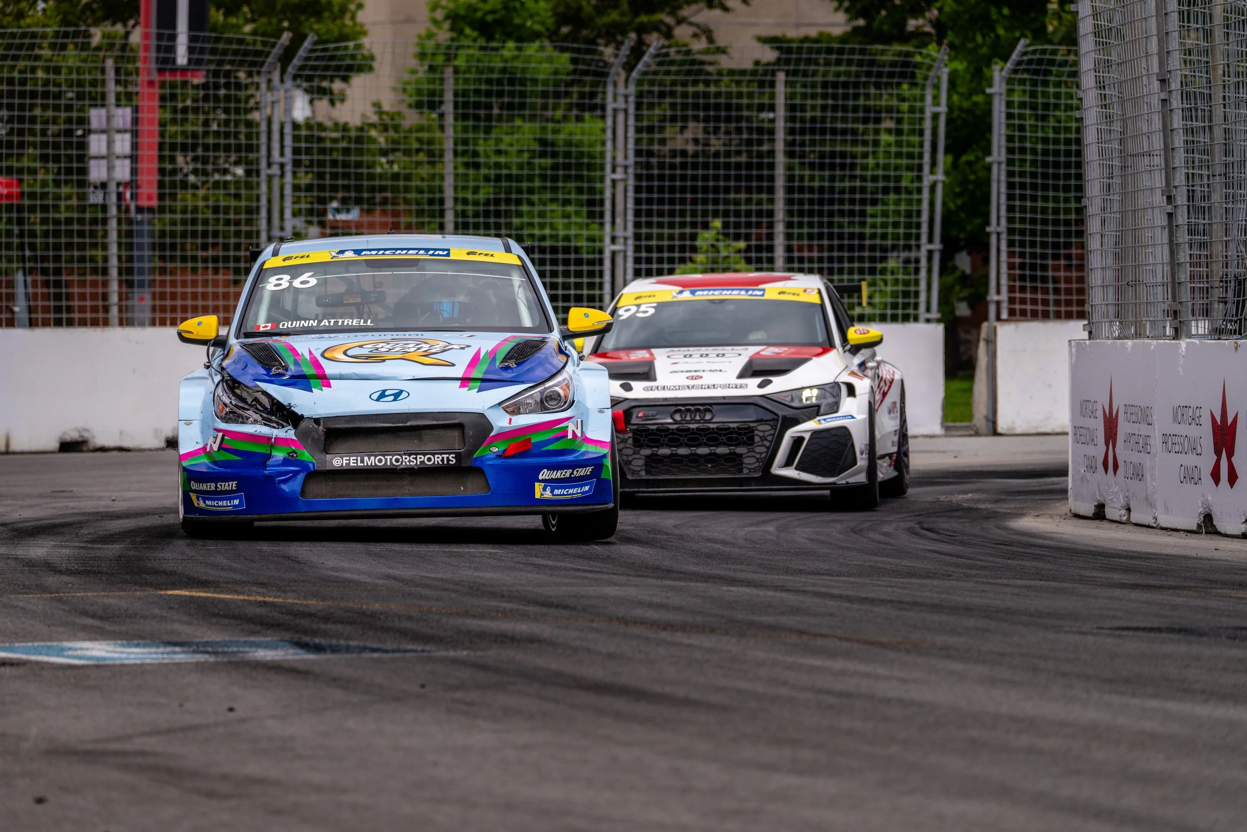 Two race cars on a track, one Hyundai with colorful livery and the number 86, and one Audi with black, white, and red design and the number 95, navigating a turn during a race.