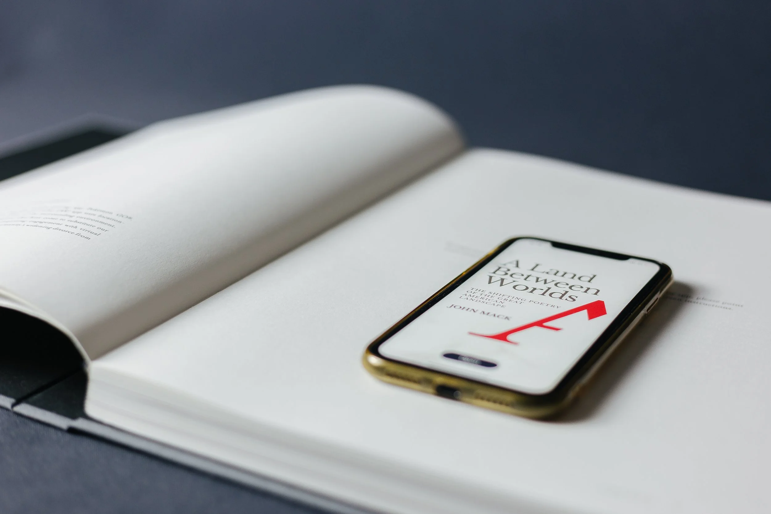 Open book with a smartphone on top displaying the cover of the book titled "A Land Between Worlds" by John Mack.