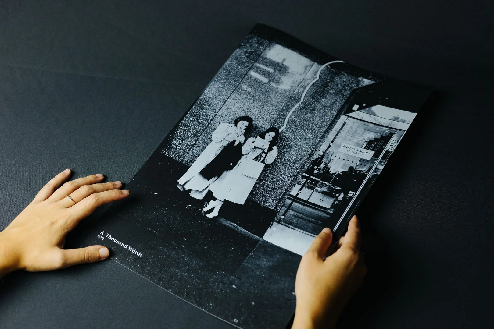 Black and white photograph of two women standing outside a building, one carrying a shopping bag, the other looking at her phone, on a dark table.
