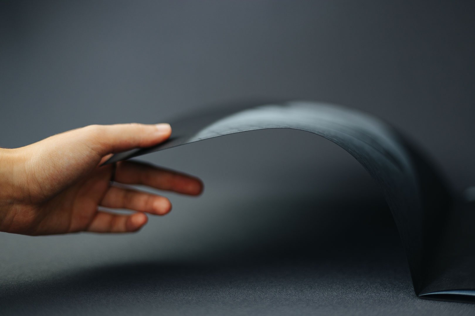 A hand lifting a corner of a thick, dark-colored paper or poster against a dark background.