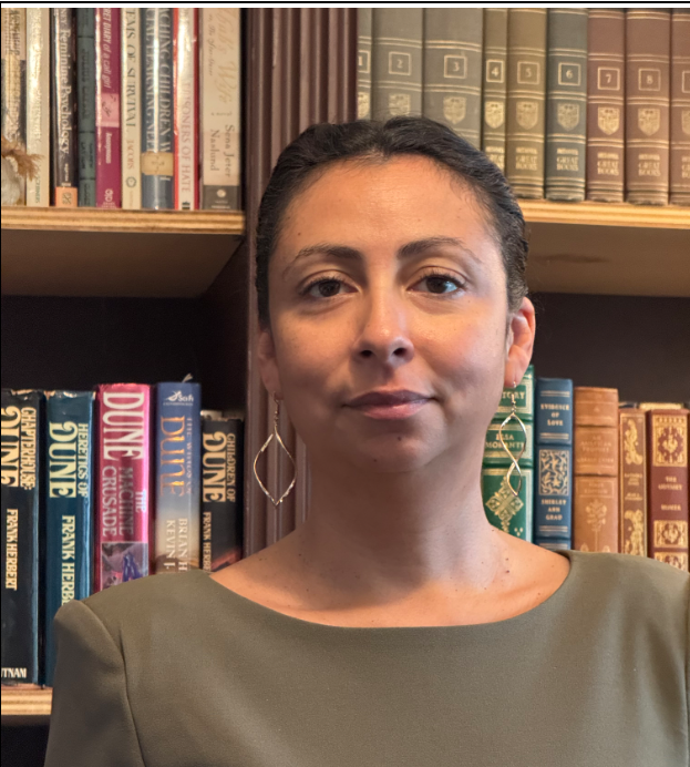 Portrait of a woman with brown hair, wearing a green top and large gold geometric earrings, standing in front of a bookshelf filled with colorful books.